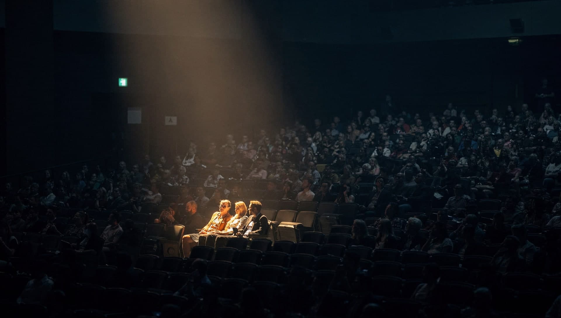 A warm shaft of overhead light falls on three people seated together in a large dark auditorium while hundreds of others around them sit in blue shadow.