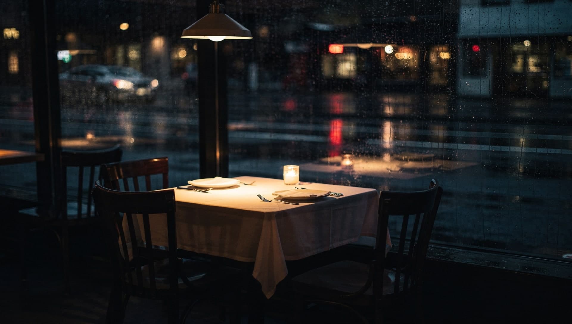 A candlelit restaurant table set with white linen and two place settings glows under a warm pendant lamp by a large rain streaked window, the rest of the room dark and empty with a wet street beyond.