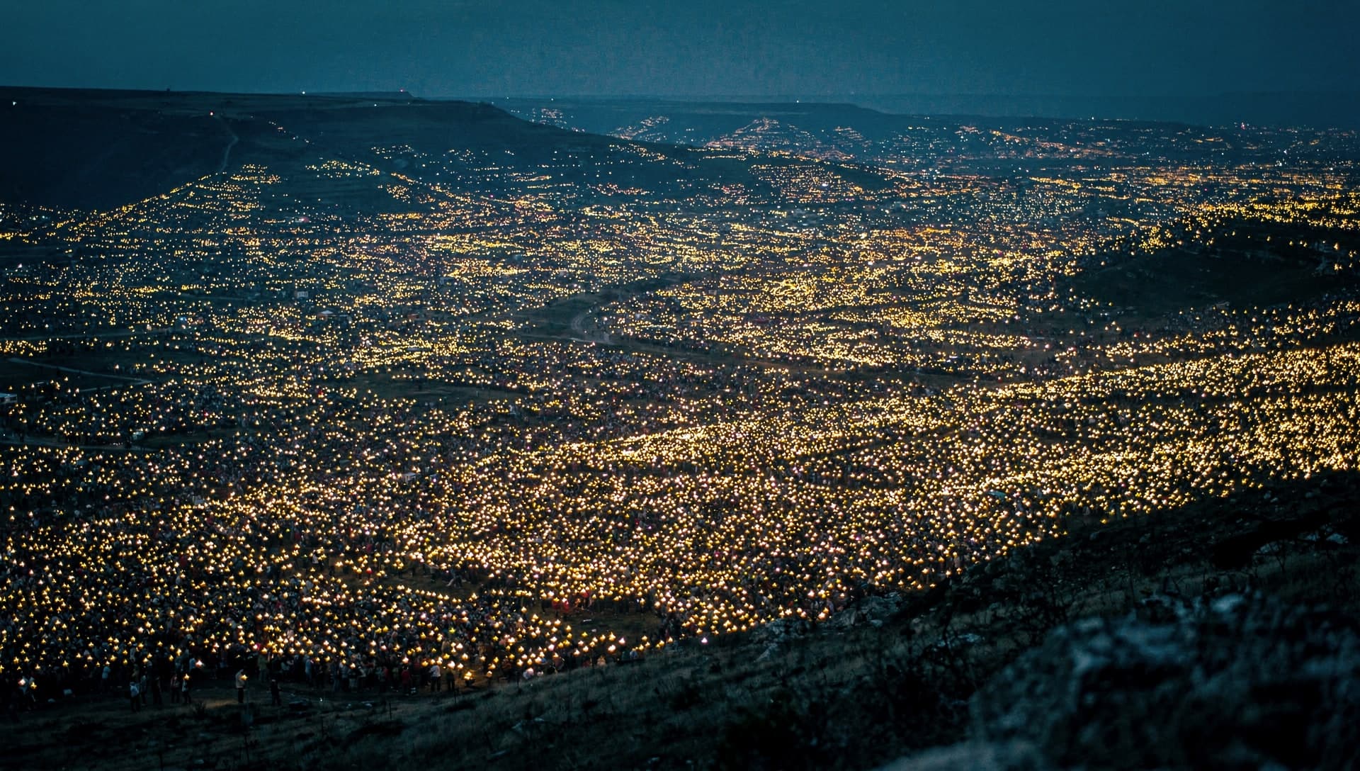 A hillside overlook at dusk above a vast valley filled to the horizon with countless tiny warm points of light from an enormous crowd holding small flames, under a deep indigo sky.