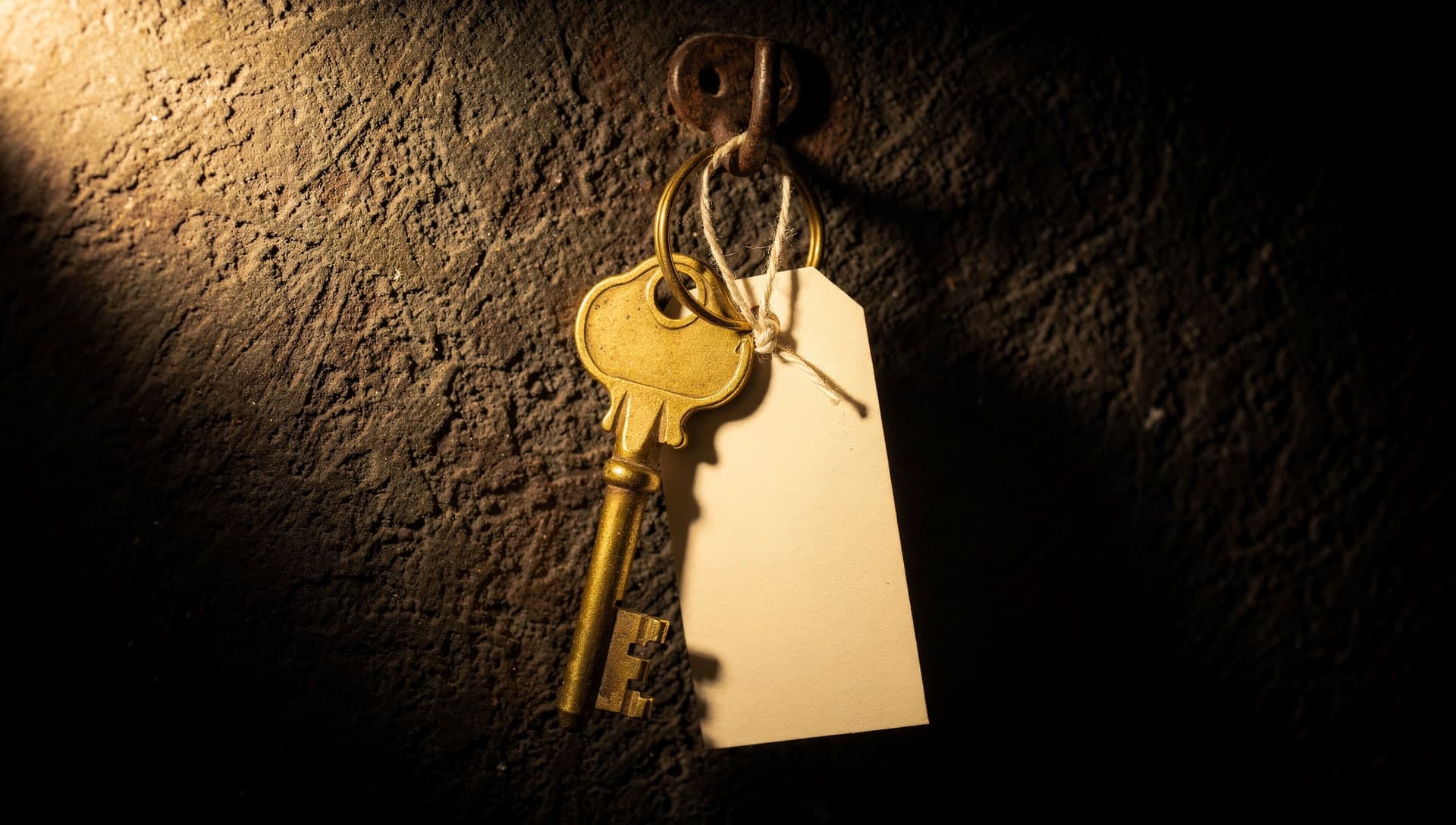 A vintage brass key on an iron hook against a dark plaster wall, a blank pale paper tag tied to it with string, lit by a single warm spotlight so the key and tag glow gold against deep shadow.
