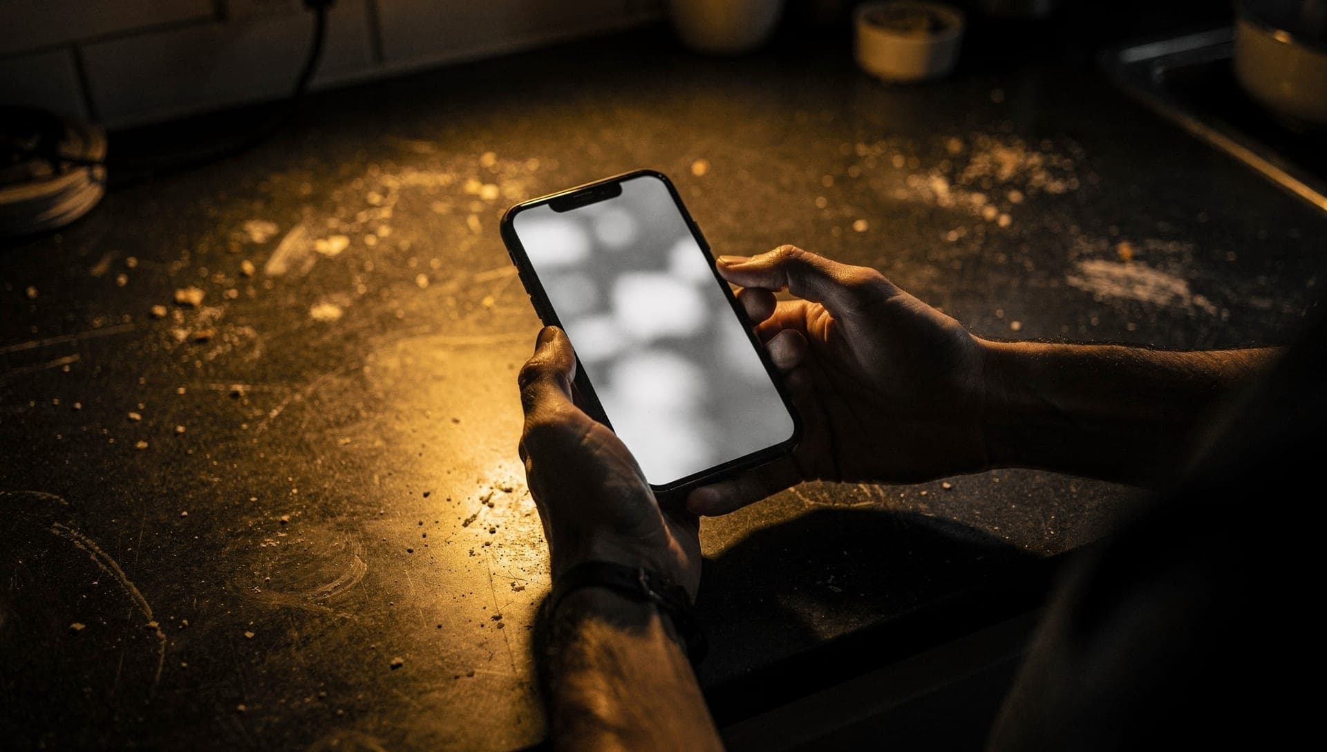 Hands holding a phone with a blurred white screen over a crumb-dusted kitchen counter in lamplight