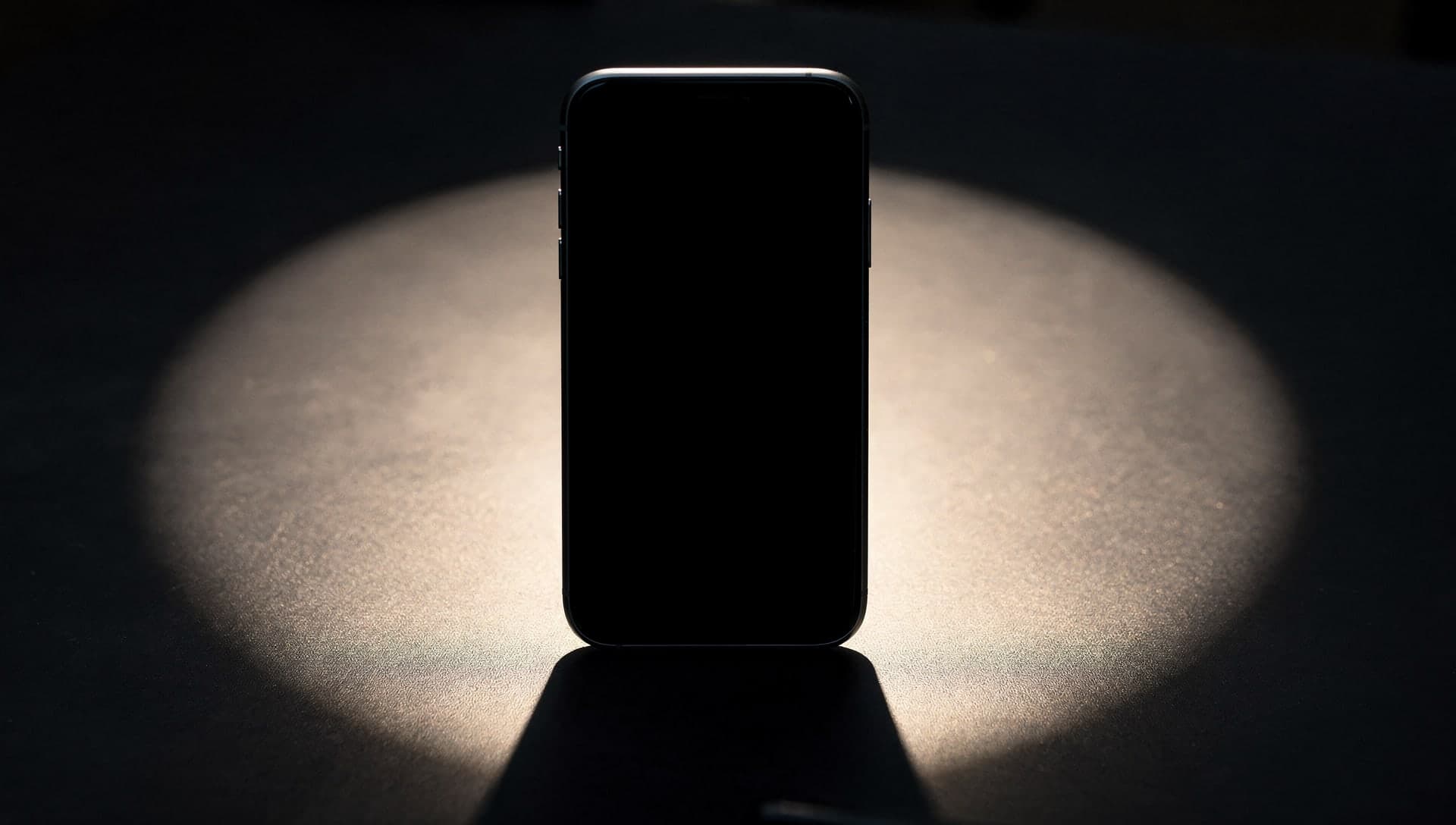 A dark smartphone standing upright in a circular pool of warm light on a dark surface