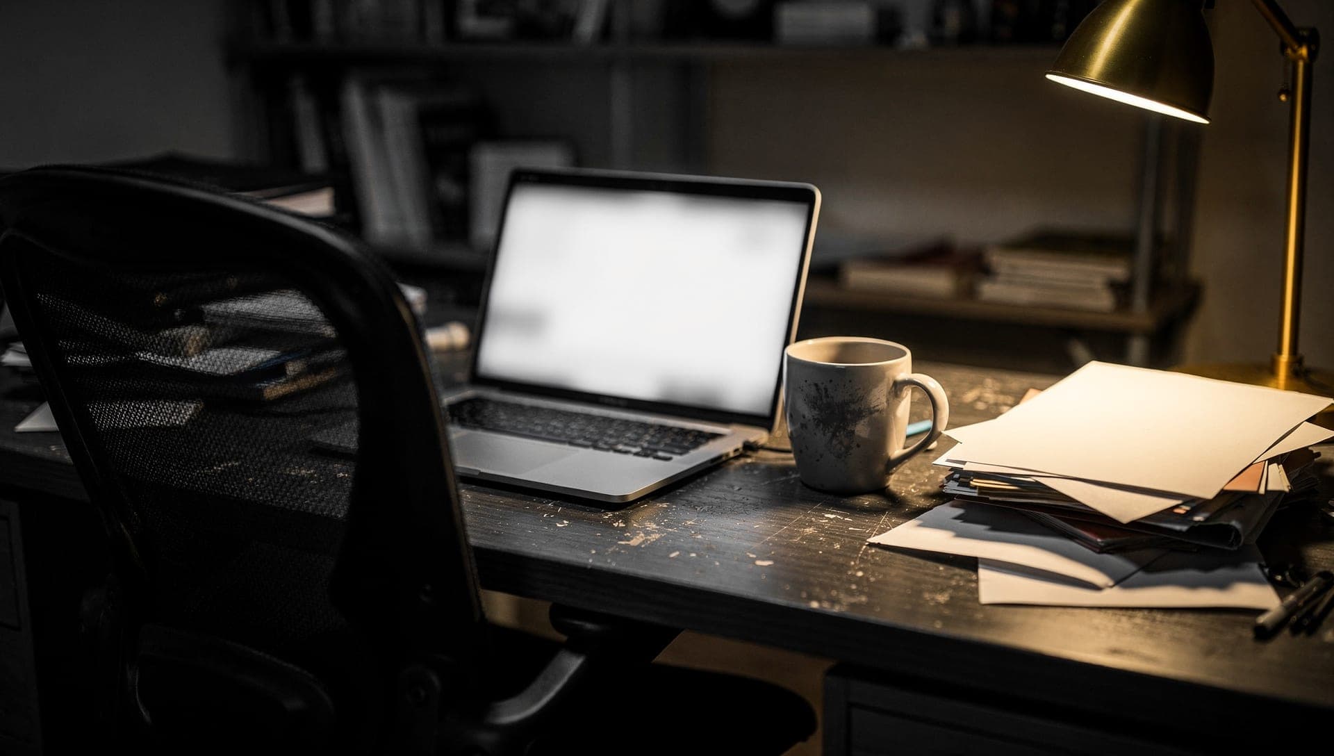 Open laptop with a blurred white screen on a scratched black desk beside a speckled mug, a stack of papers, and a brass lamp