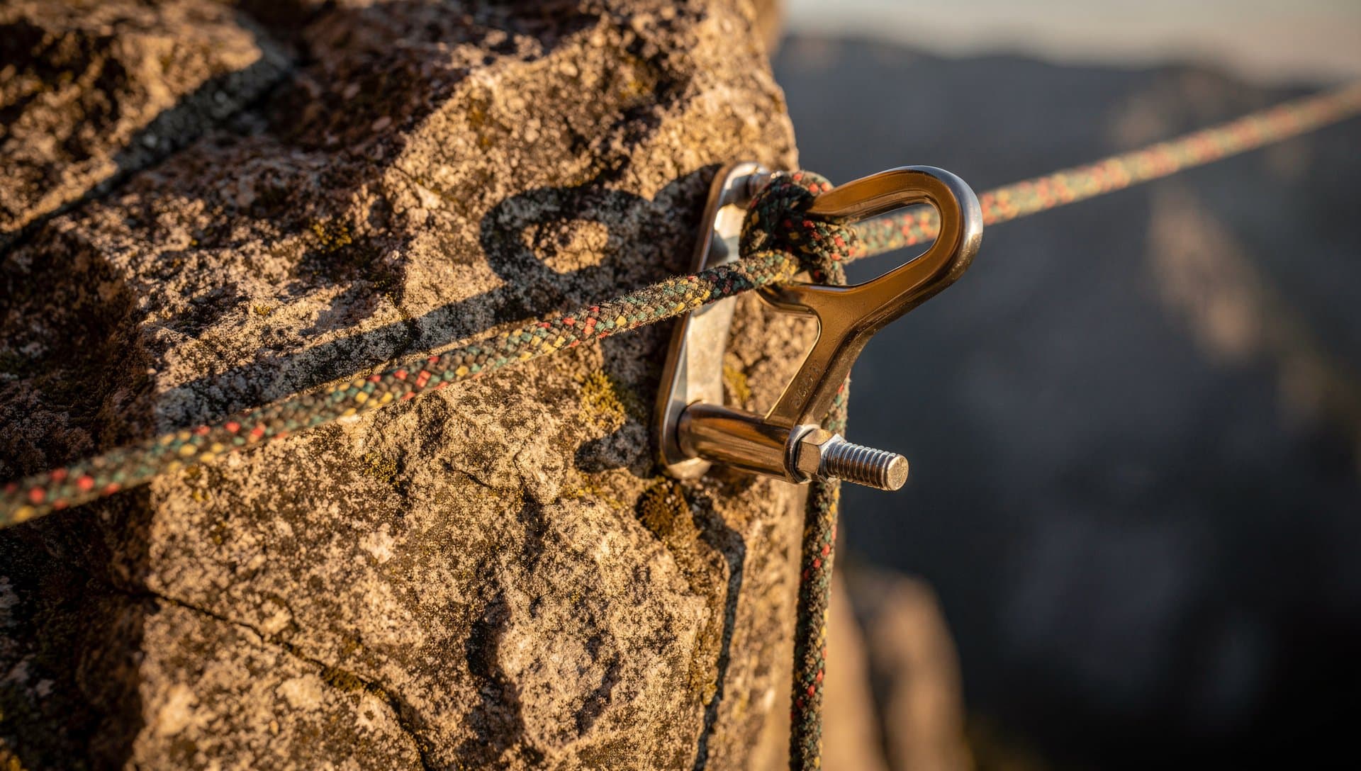 A climbing rope tied through a stainless steel ring anchor bolted into a sun-warmed rock face in warm afternoon light, a canyon blurred behind