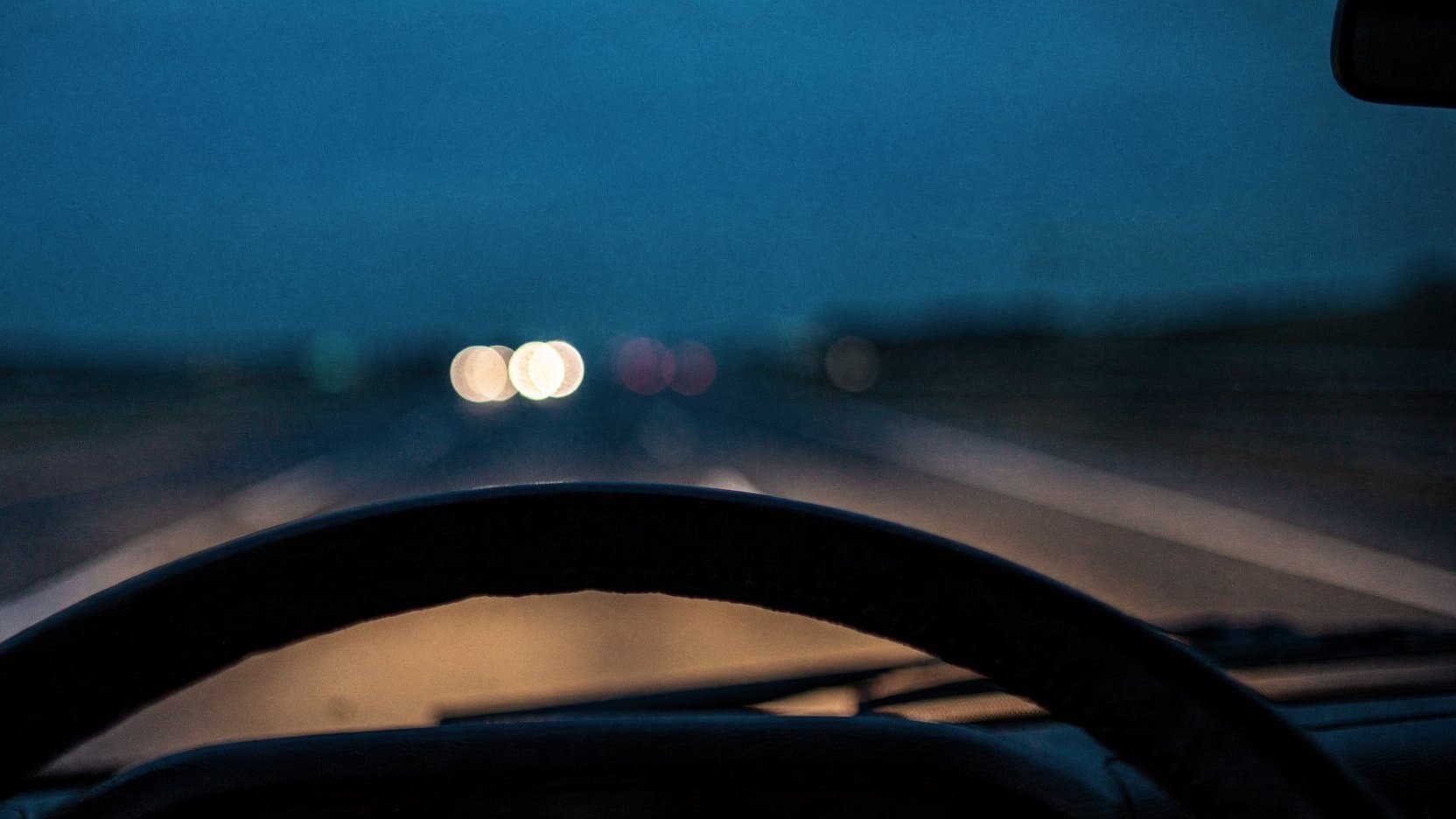 The driver's view over a dark steering-wheel rim at dusk, an open road blurring ahead through the windshield with warm bokeh headlights