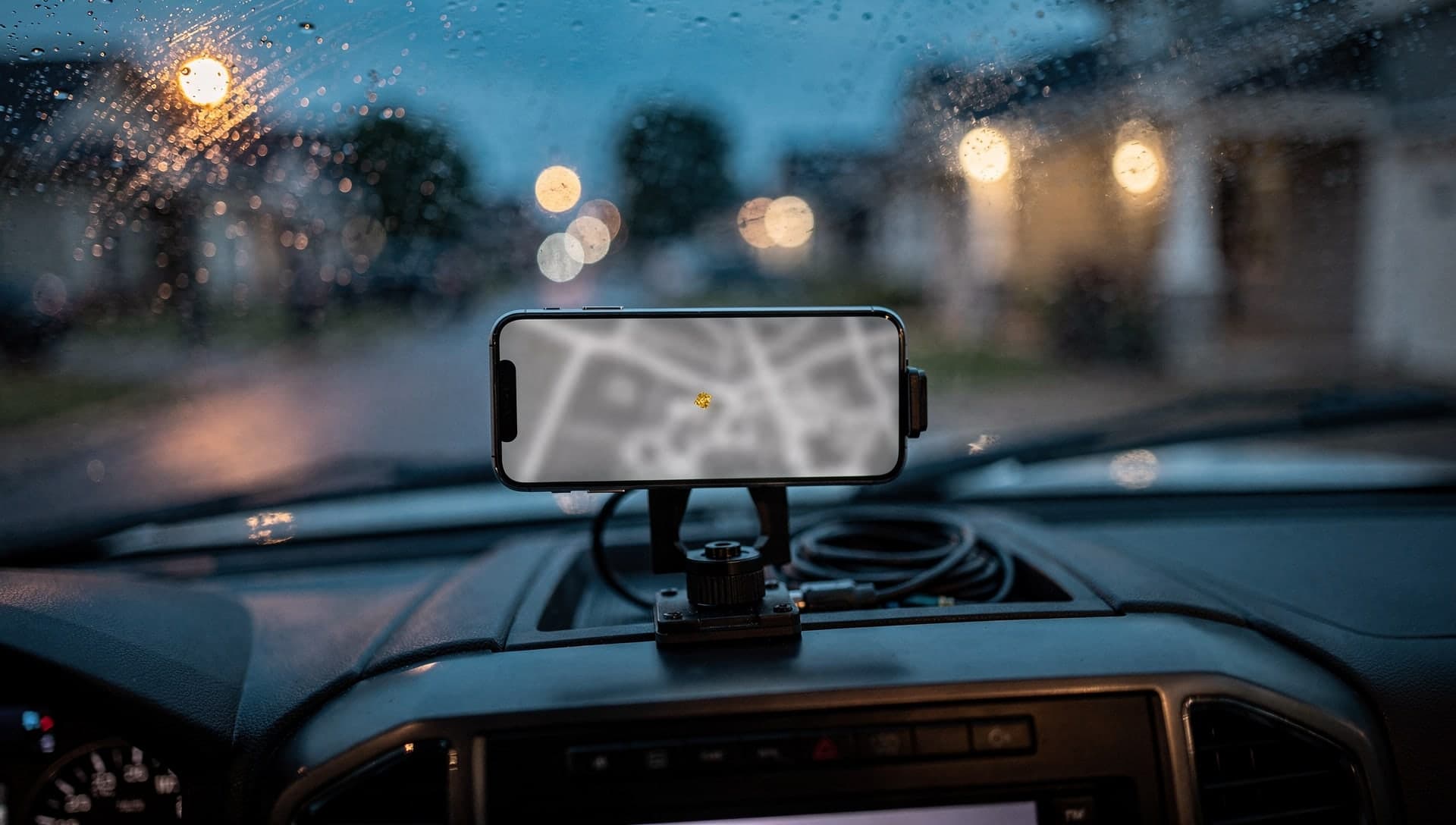 A dash-mounted phone showing a blurred map with a gold pin through a rainy windshield at dusk