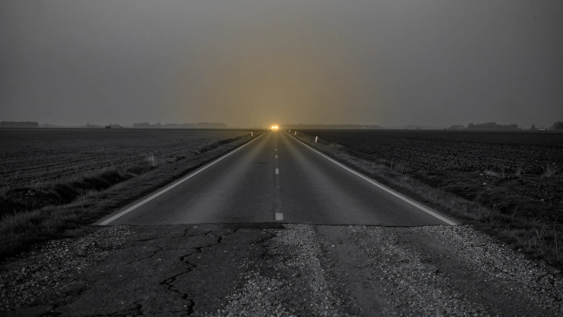 Empty two-lane road running toward one pair of distant headlights across flat fields at dusk