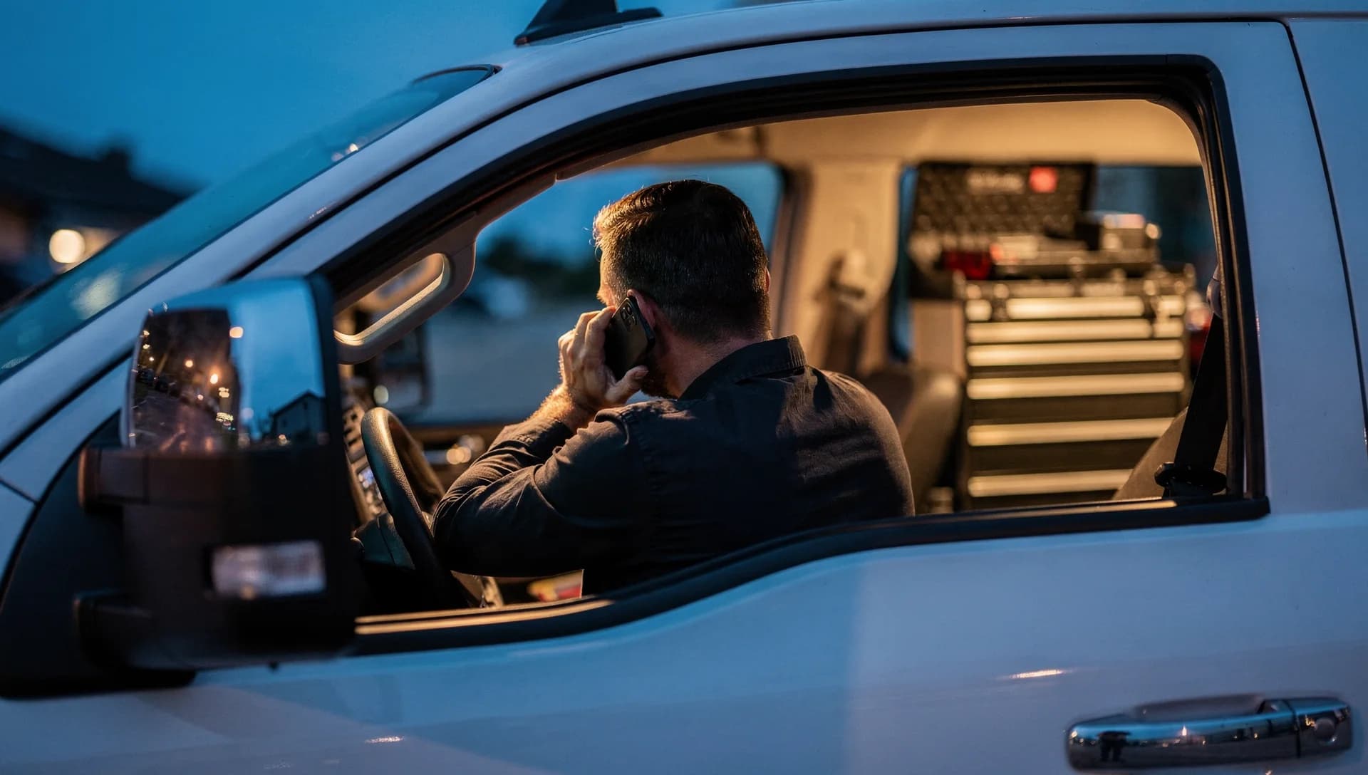 A contractor seen from behind taking a phone call in his truck cab at dusk