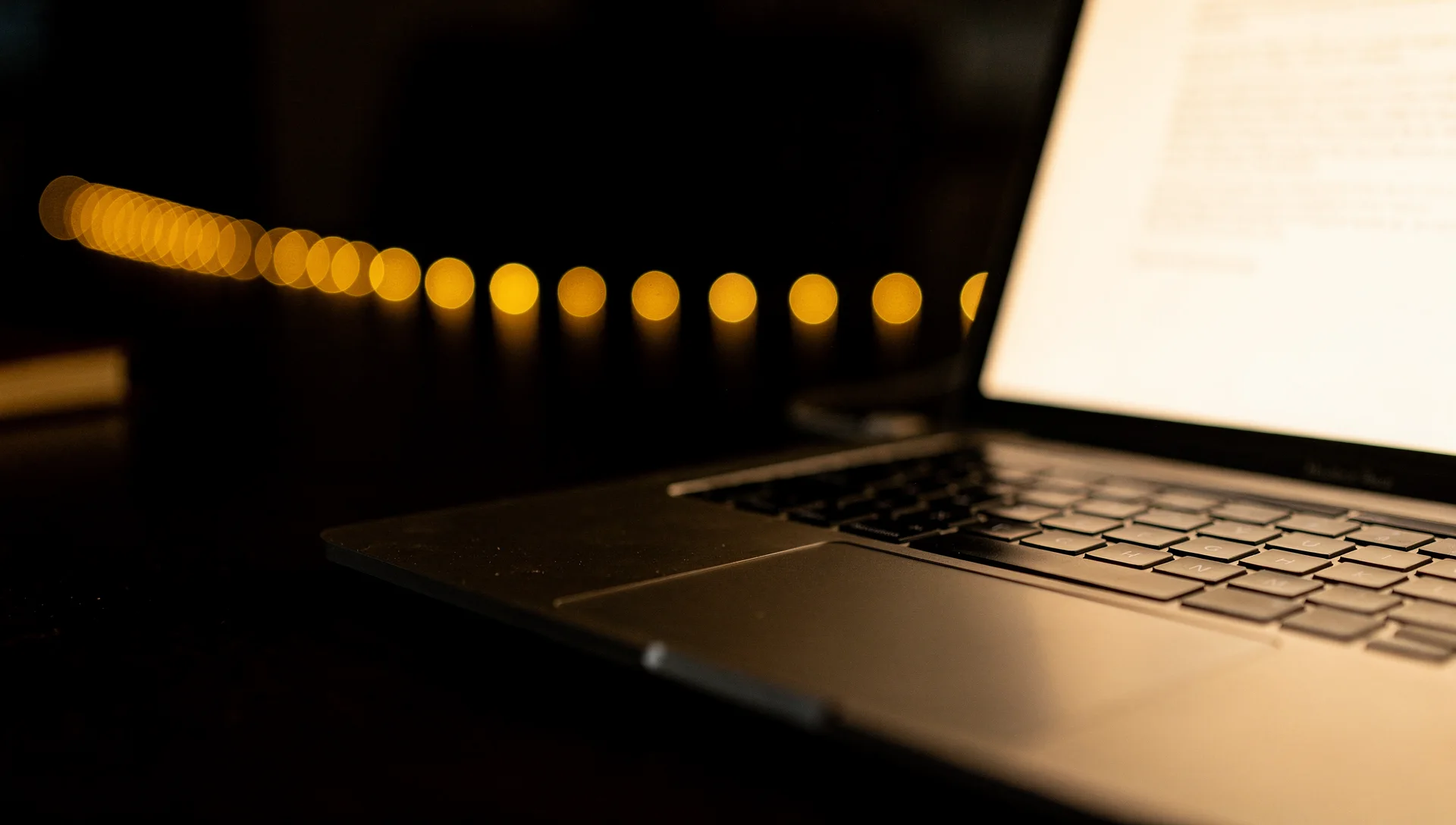 A laptop screen glowing with blurred text beside a string of gold bokeh lights in the dark