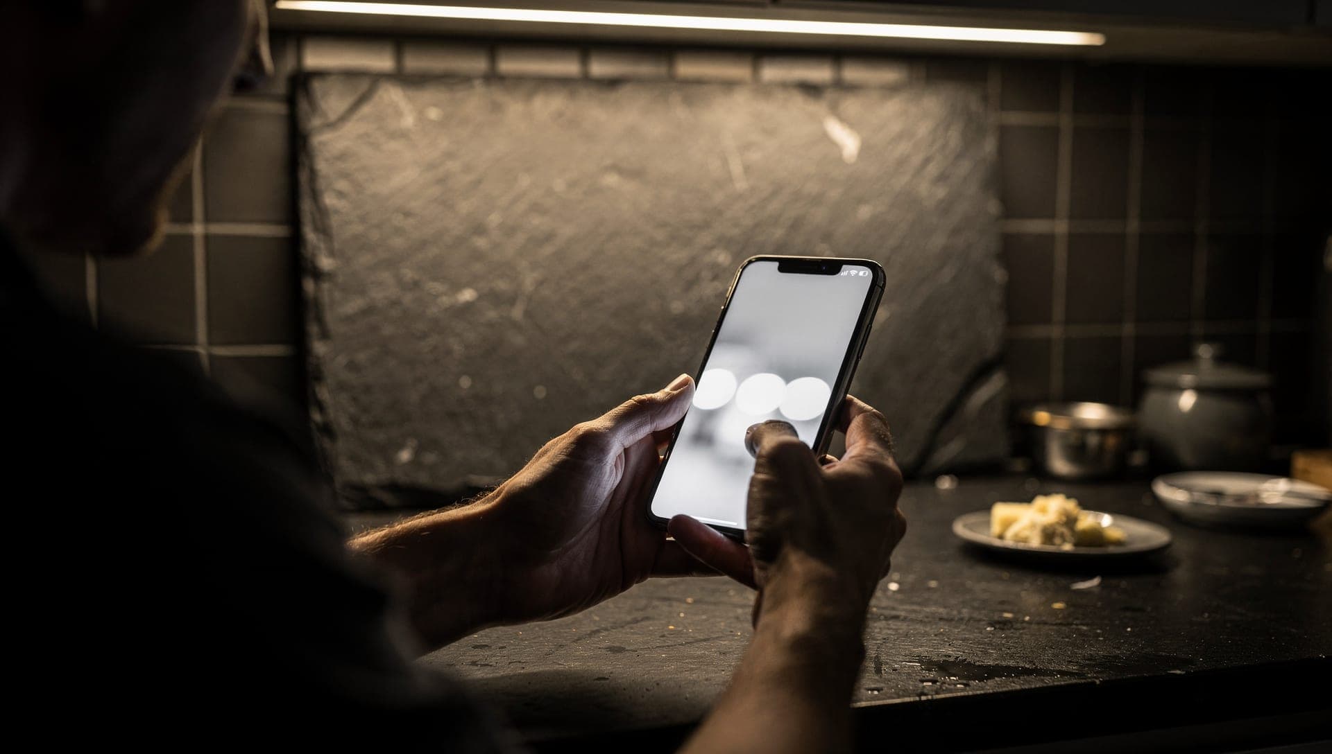 Over-the-shoulder view of hands holding a phone with a blurred white screen at a dark kitchen counter under a strip light
