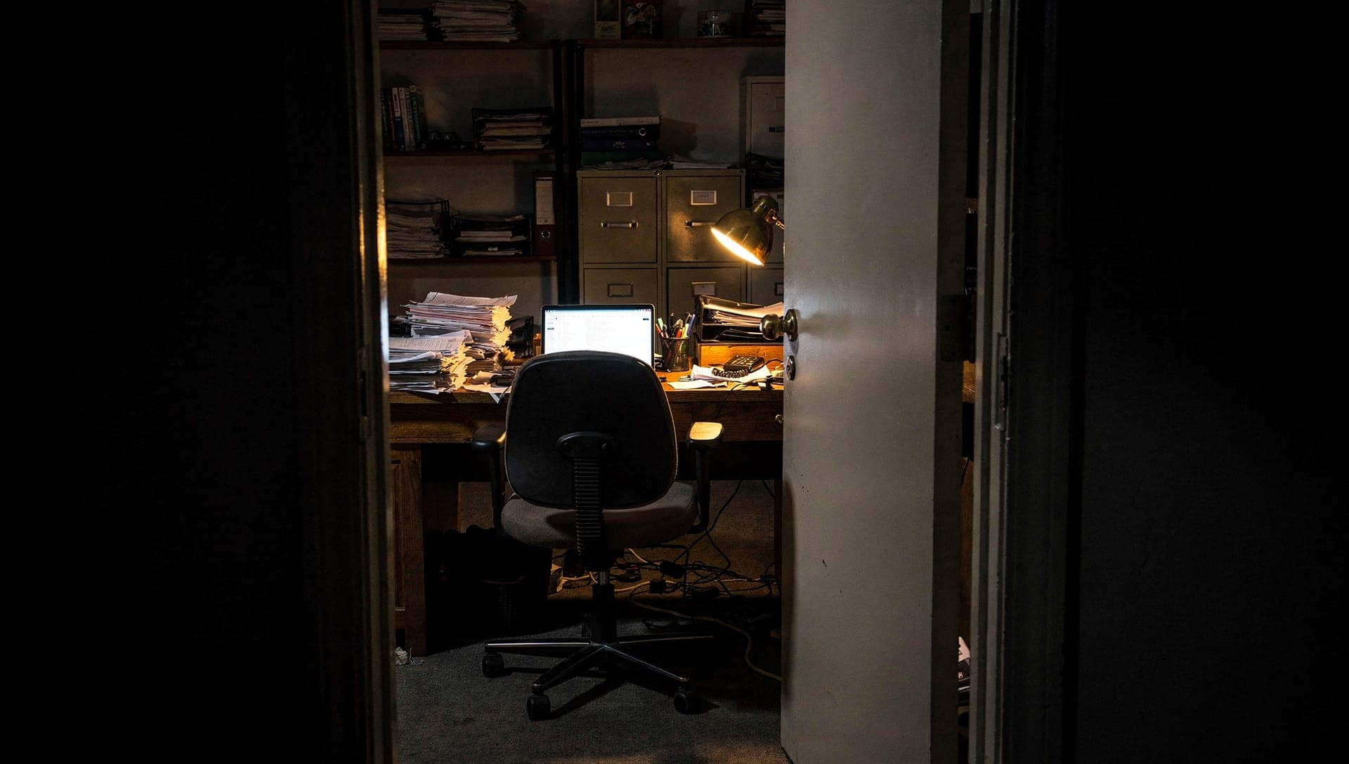 Open doorway into a cluttered back office at night, an empty desk chair in front of a glowing laptop, paper stacks, file cabinets, and one desk lamp