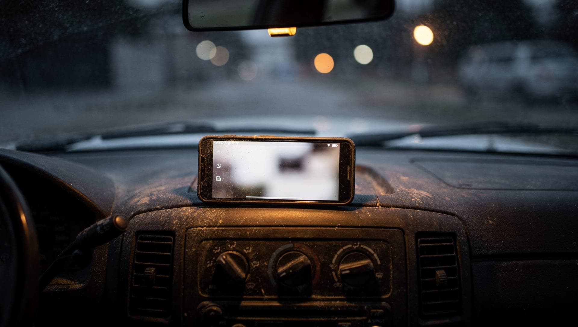 Phone propped on a dusty van dashboard with a blurred screen, rain on the windshield and blurred street lights beyond