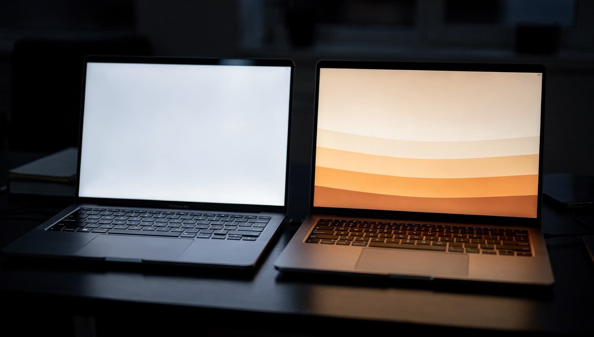 Two laptops side by side in the dark, one screen blank white and one glowing with a warm gradient