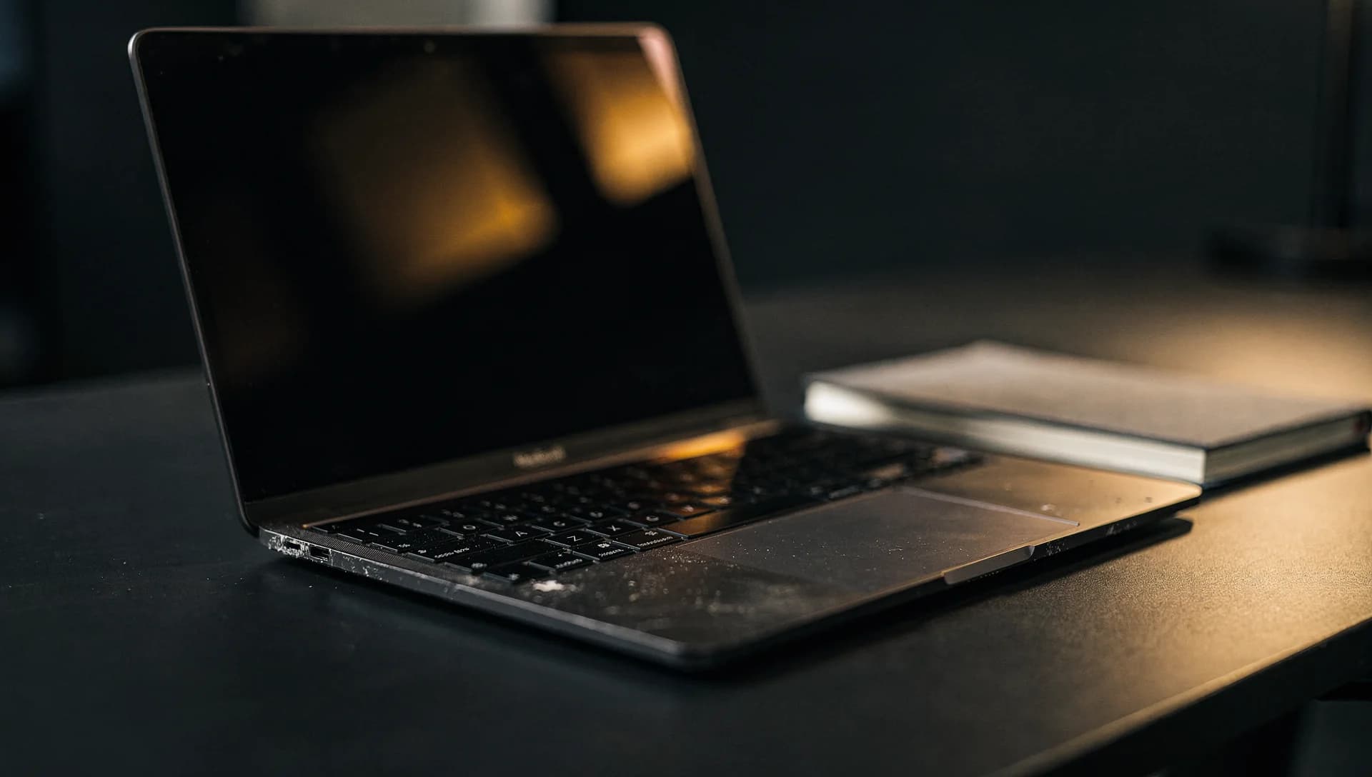 An open laptop with a dark reflective screen beside a notebook on a dark desk