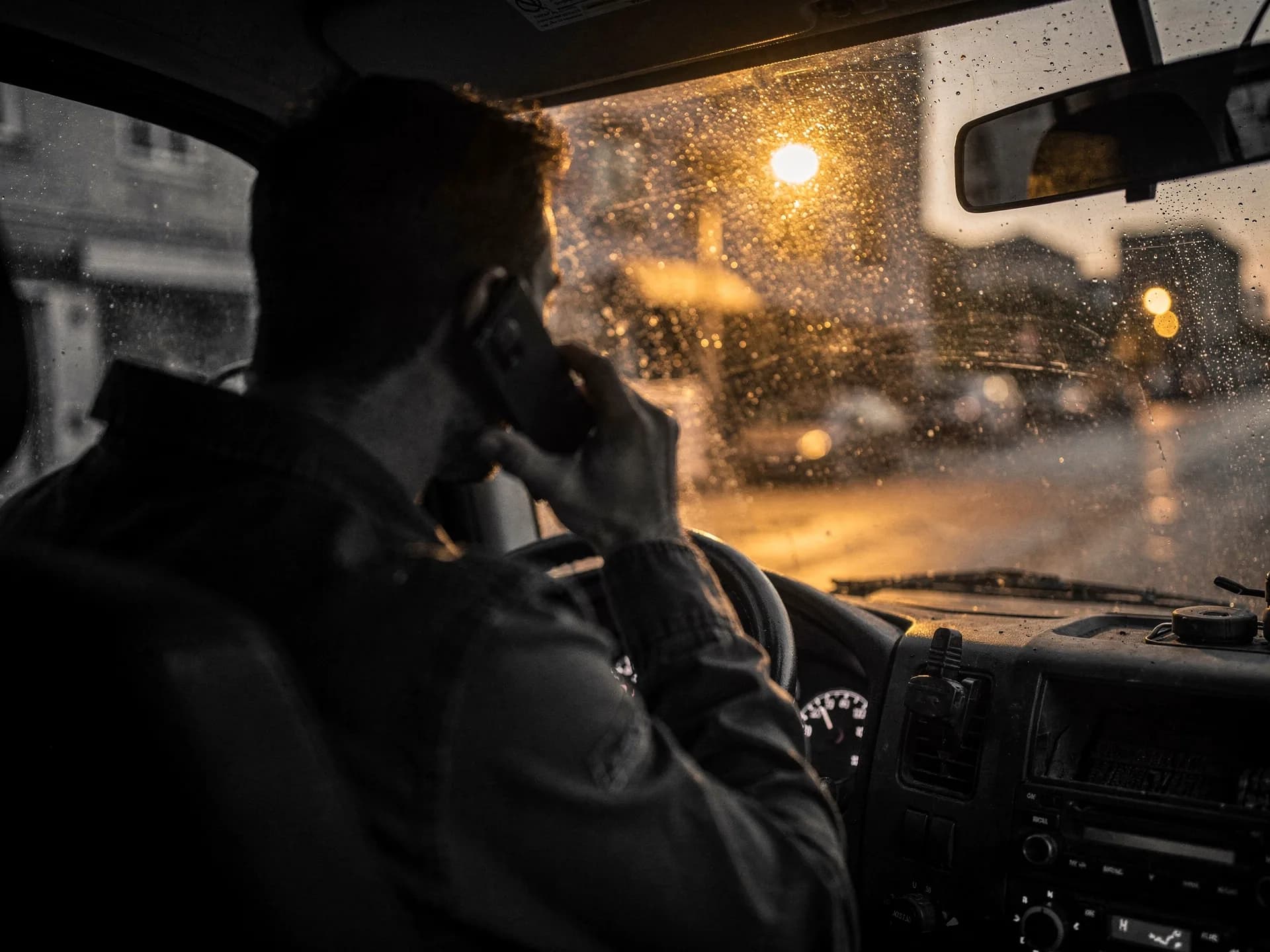 A man seen from behind holding a phone to his ear in a work truck cab, a warm streetlight blooming through the rain-speckled windshield