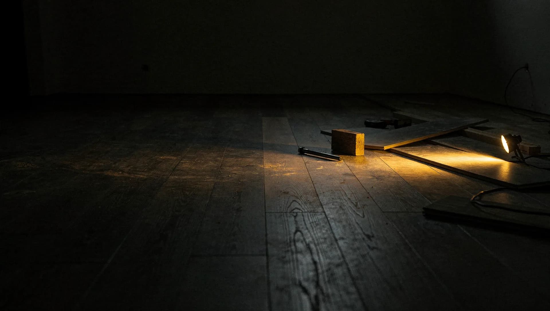 A gold work light rakes across a half-laid run of plank flooring at night, a tapping block and pull bar beside loose boards, the seams catching the light, the left side falling into shadow.