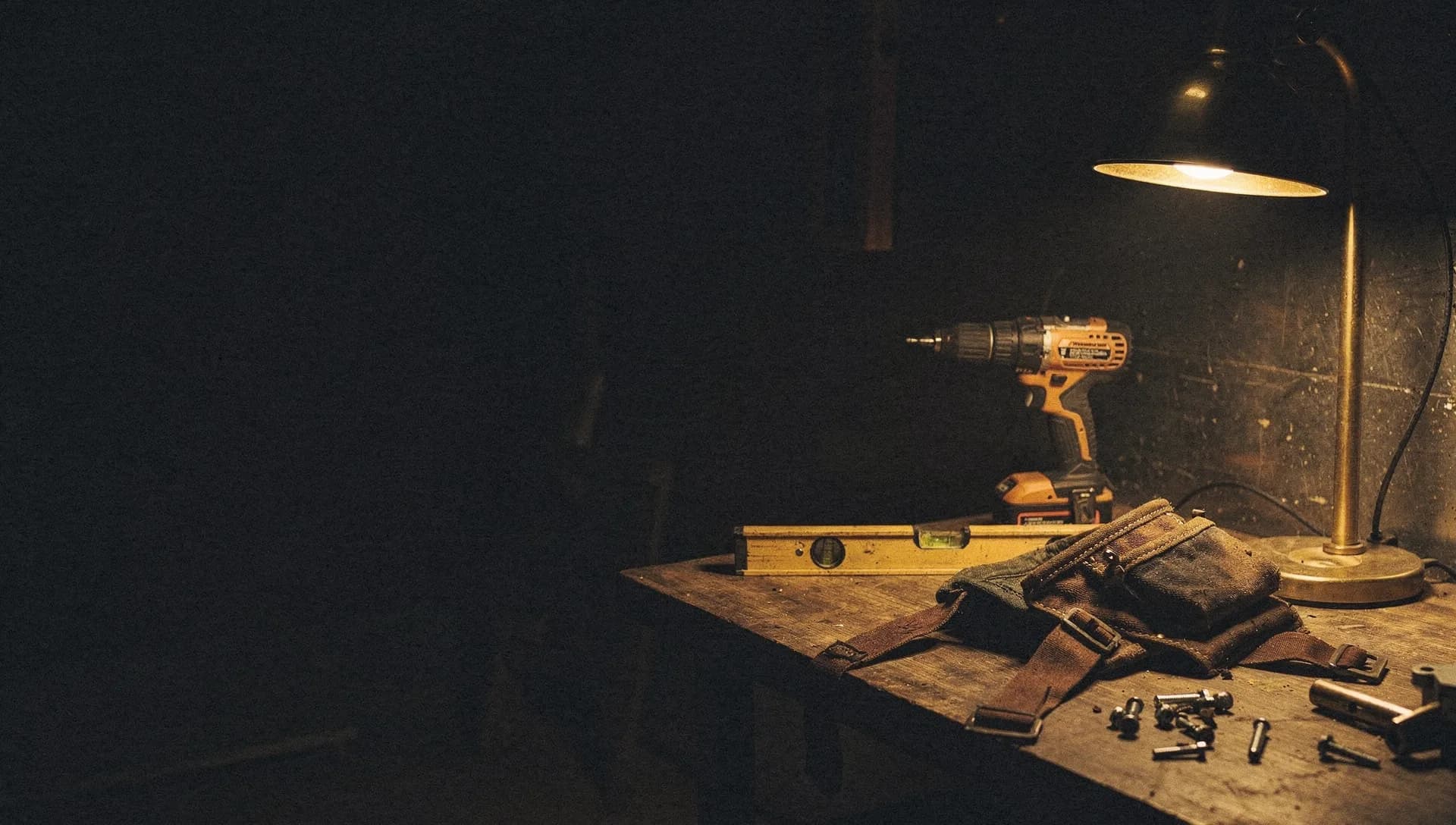 A gold work light catches a cordless drill, a level, and a tool belt laid across a wooden workbench at night, a few fasteners scattered nearby, the left side falling into shadow.