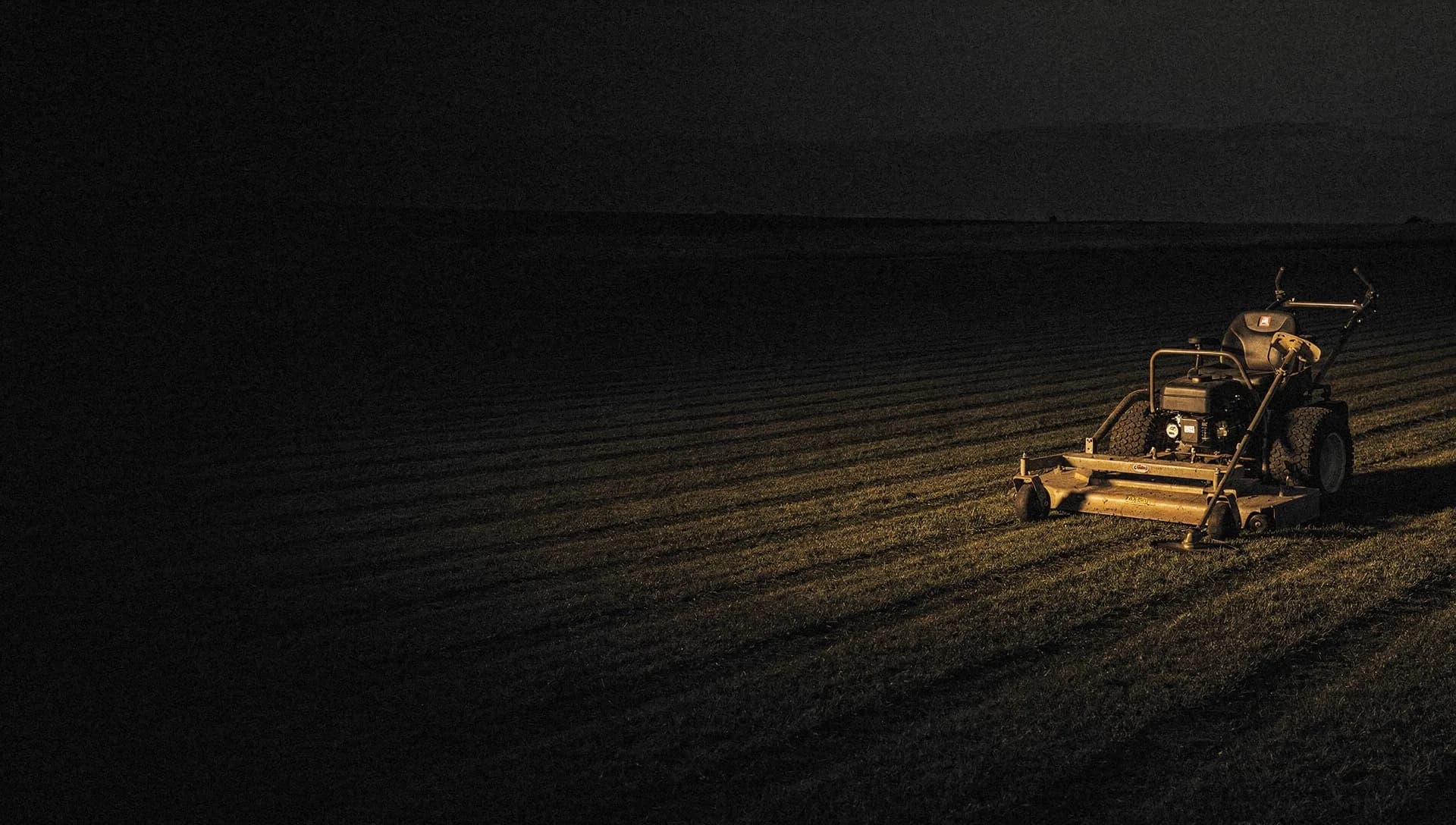 At dusk a gold light rakes across freshly striped turf and the deck of a mower with a trimmer leaned against a tailgate, the left side of the lawn falling into shadow.