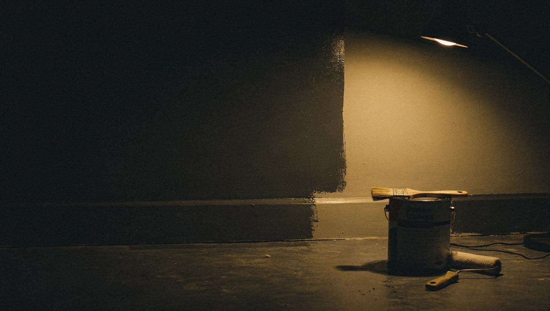 A gold work light catches a crisp painted cut line along trim at night, a brush laid across an open can and a roller in a tray nearby, the left side of the room falling into shadow.