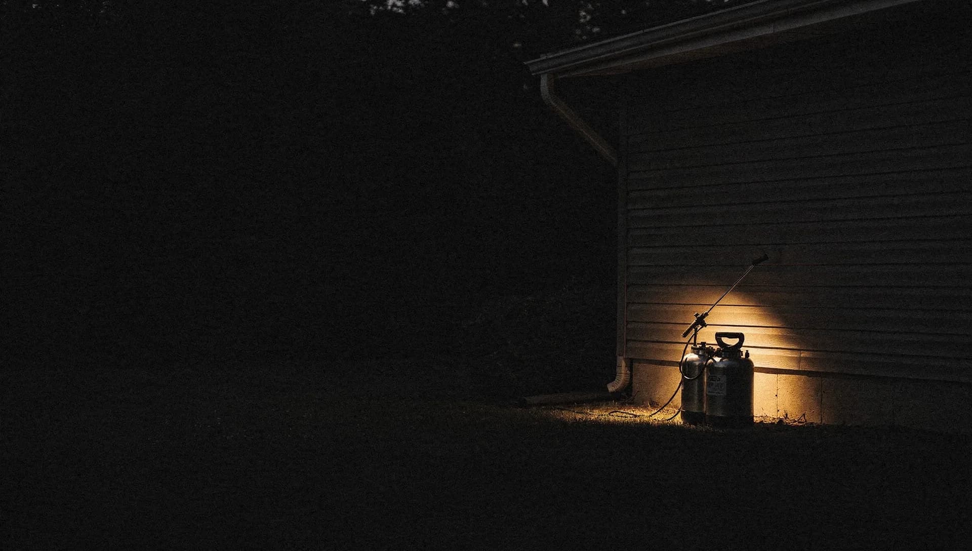 At dusk a gold light catches a pest control sprayer wand and tank resting against a home's foundation, the dark eave and corner behind, the left side falling into shadow.