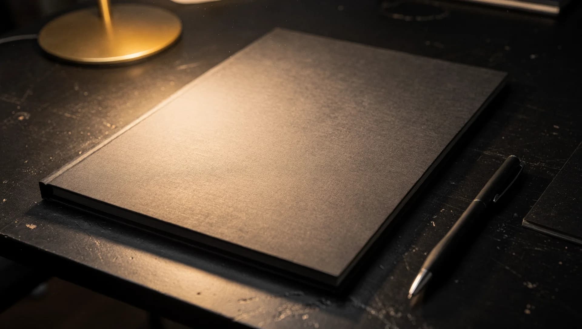 A closed graphite document folder beside a pen under a warm desk lamp