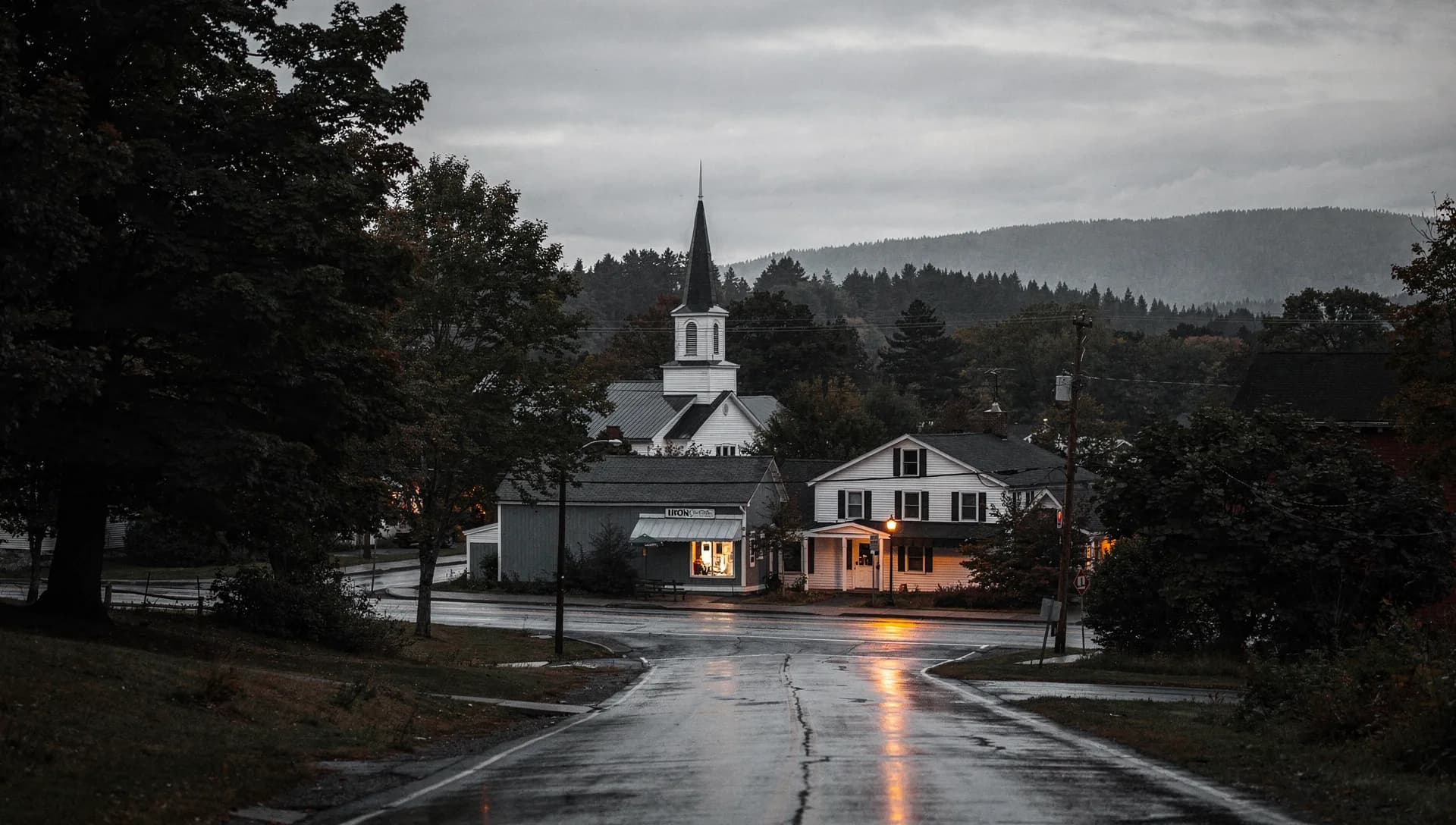 A wet small-town main street at dusk leading to a white church steeple, a lit corner storefront and a farmhouse with a glowing porch light under pine ridges