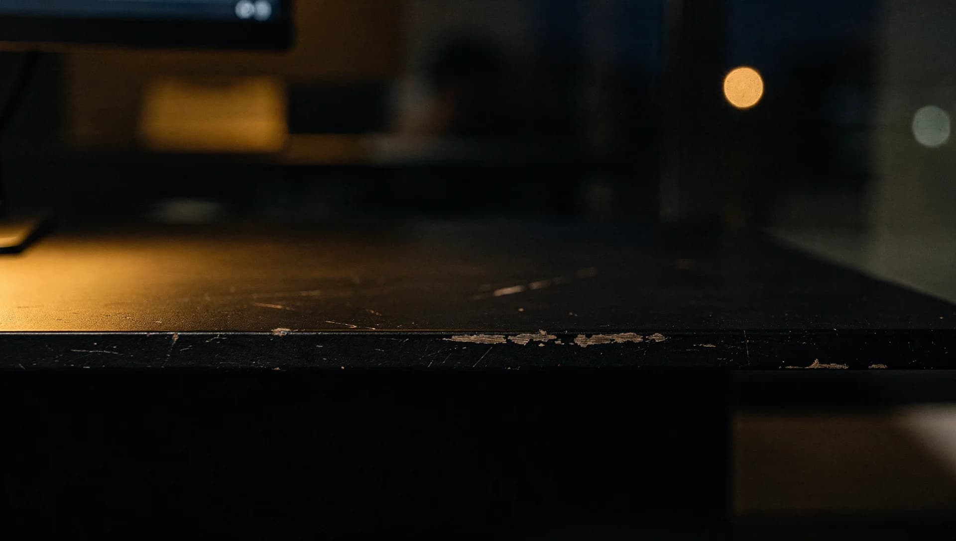 Extreme close-up of a scratched matte-black desk edge with a monitor corner and warm bokeh