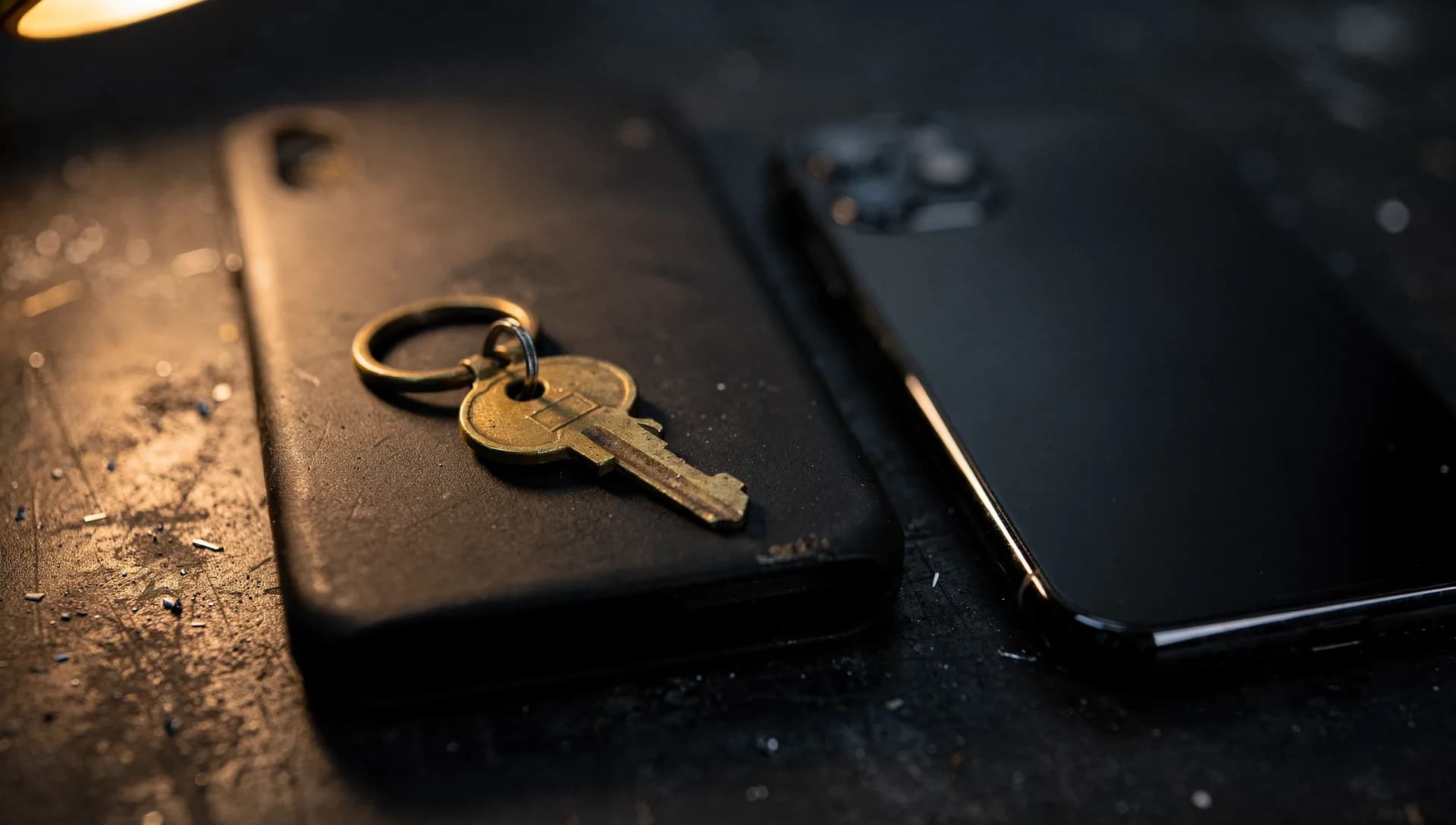 A brass key on a ring resting on a black phone beside another phone on a dark bench