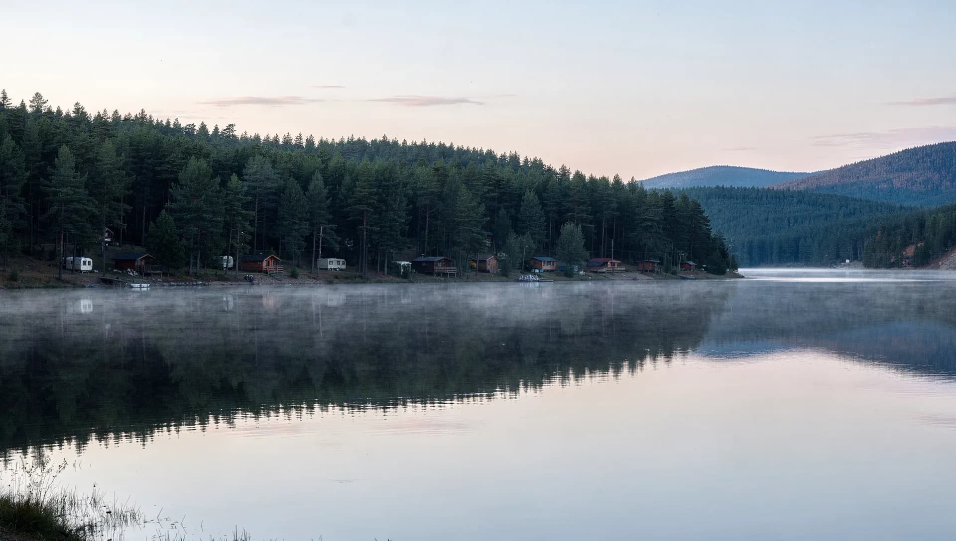 A still lake at dawn with mist rising off the surface, a forested shoreline of dark pines dotted with small cabins and campers reflected in the water, wooded hills fading into a pink sky