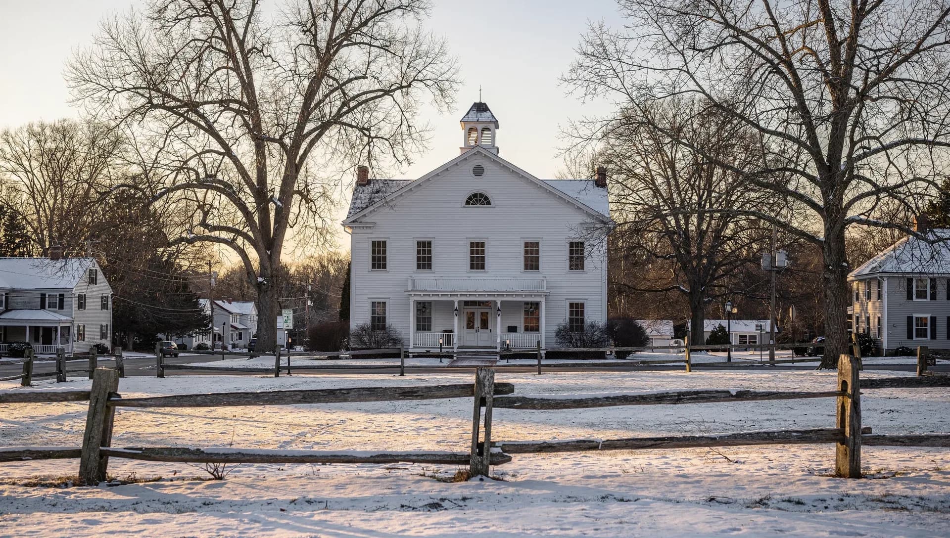 A white Greek Revival meetinghouse with a cupola and columned porch on a snow-dusted village green, a weathered split-rail fence in the foreground and bare trees glowing in low winter sun