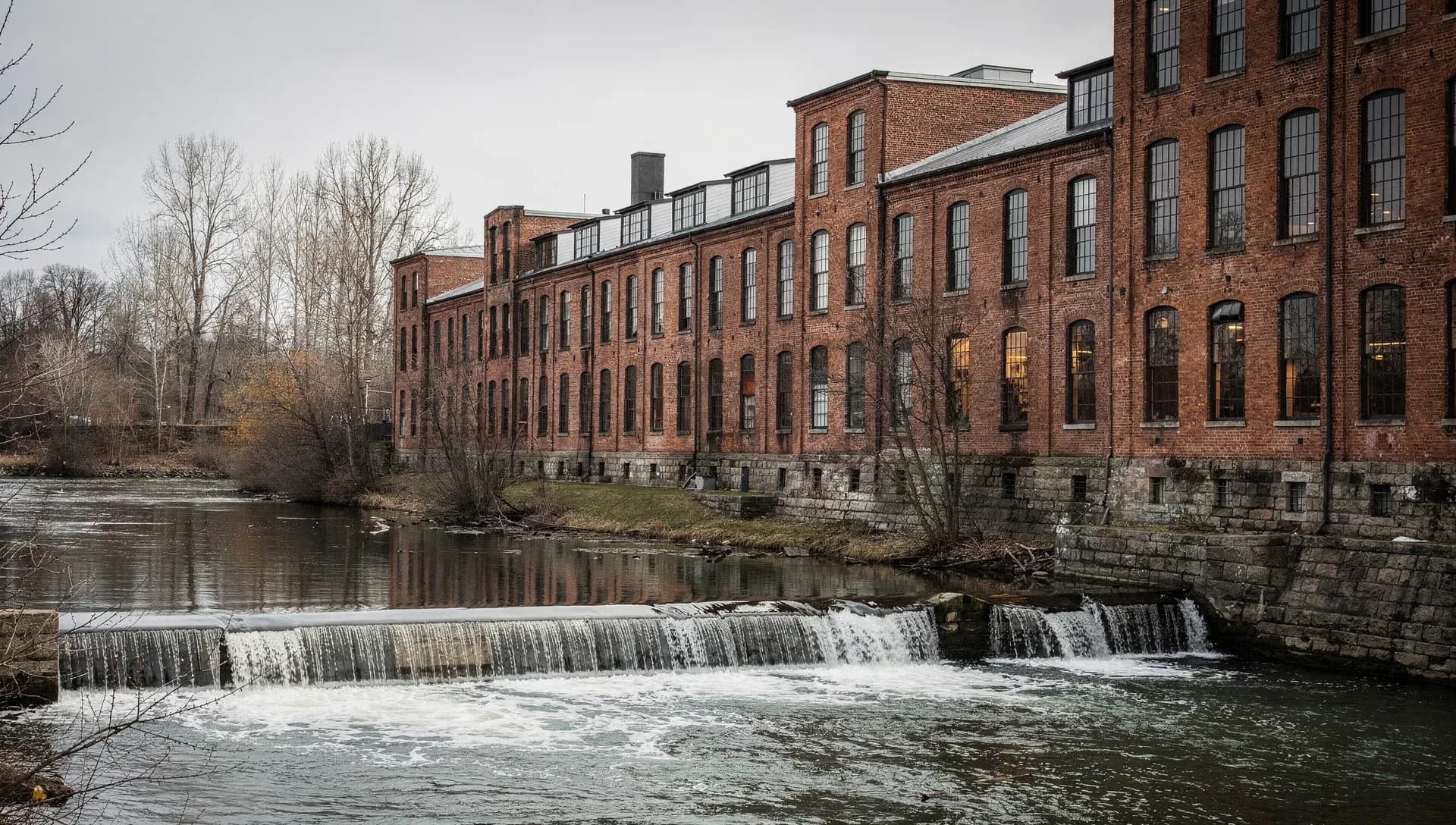 A long red-brick mill building with rows of tall windows running along a river, a low dam spilling white water across the foreground under a grey sky, bare trees on the far bank