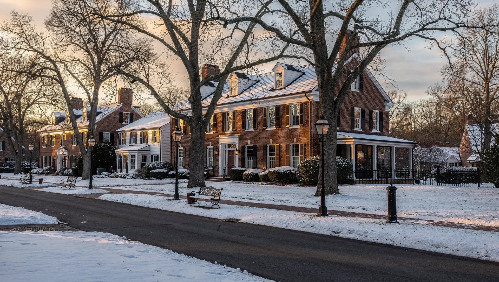 A snow-covered tree-lined residential street at sunrise with stately brick and white colonial homes, wrought-iron benches and black lampposts along the sidewalk beneath bare oaks