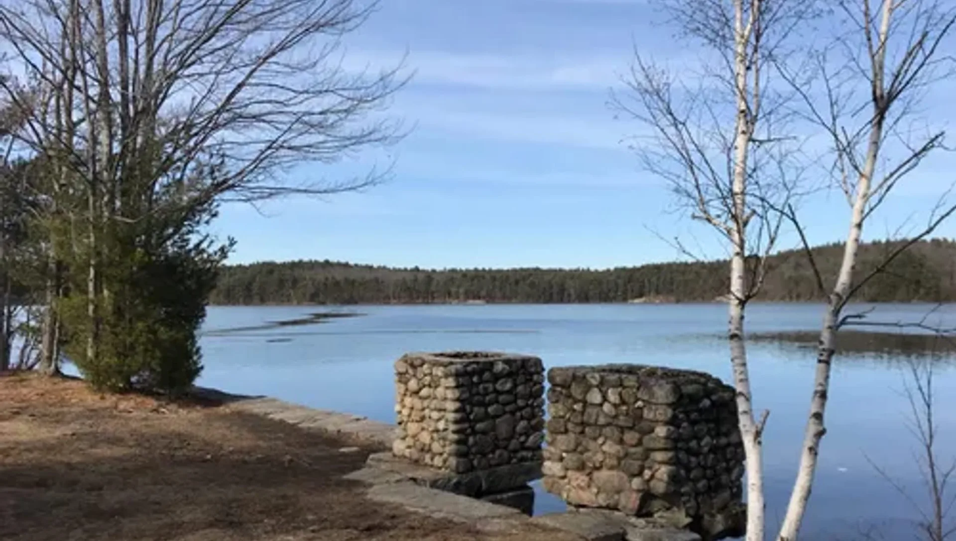 A calm lake under a pale blue sky seen between bare birches, two old cobblestone gate pillars standing at the wooded shoreline
