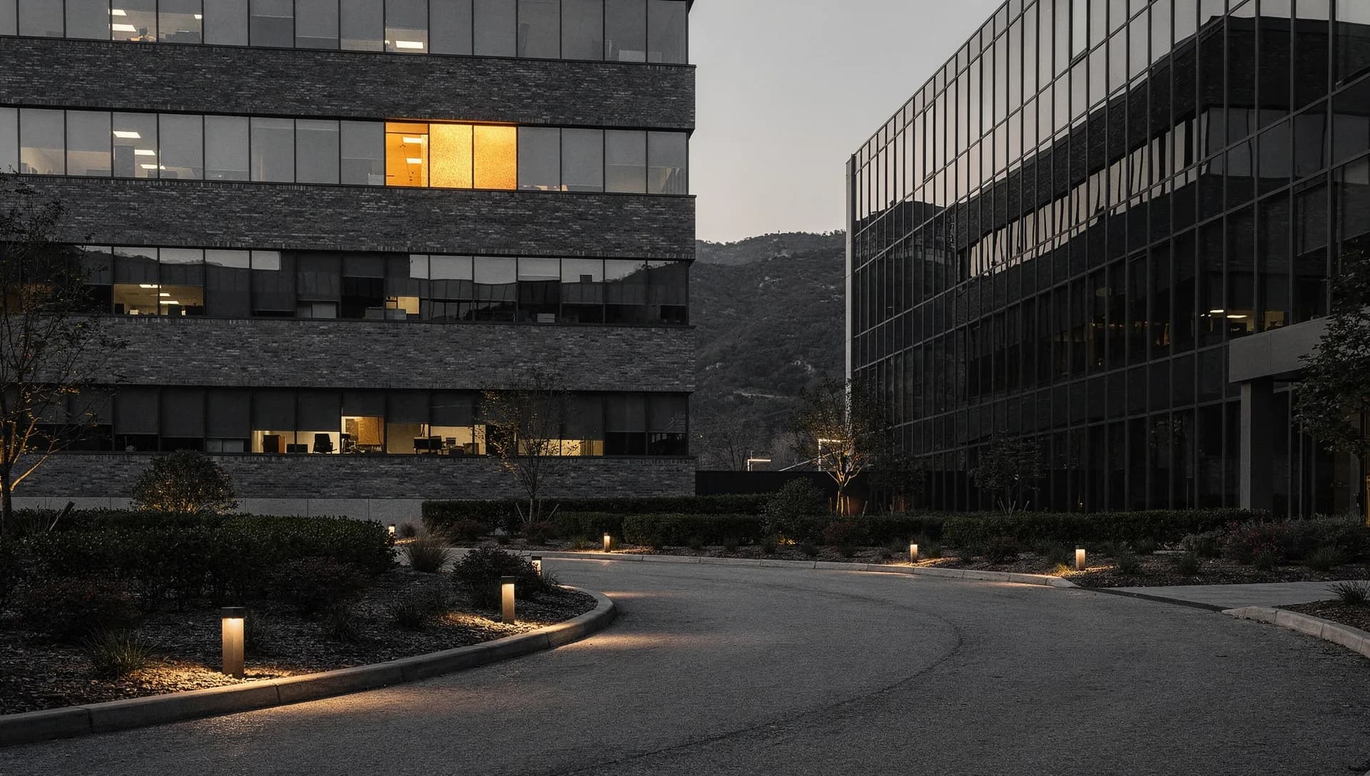 A curved office-park drive at dusk between a brick office block with one lit gold window and a dark glass building, low path lights along trimmed hedges