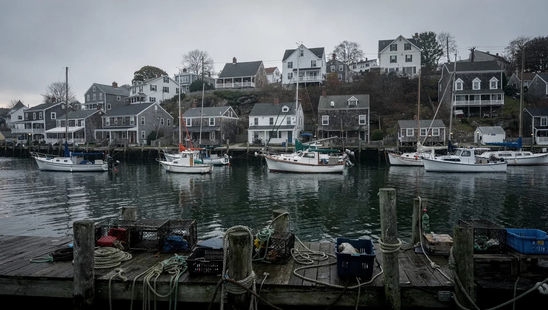 A calm grey harbor lined with moored fishing boats and sailboats, shingled houses climbing the hillside behind, a weathered dock stacked with lobster traps and coiled rope in the foreground