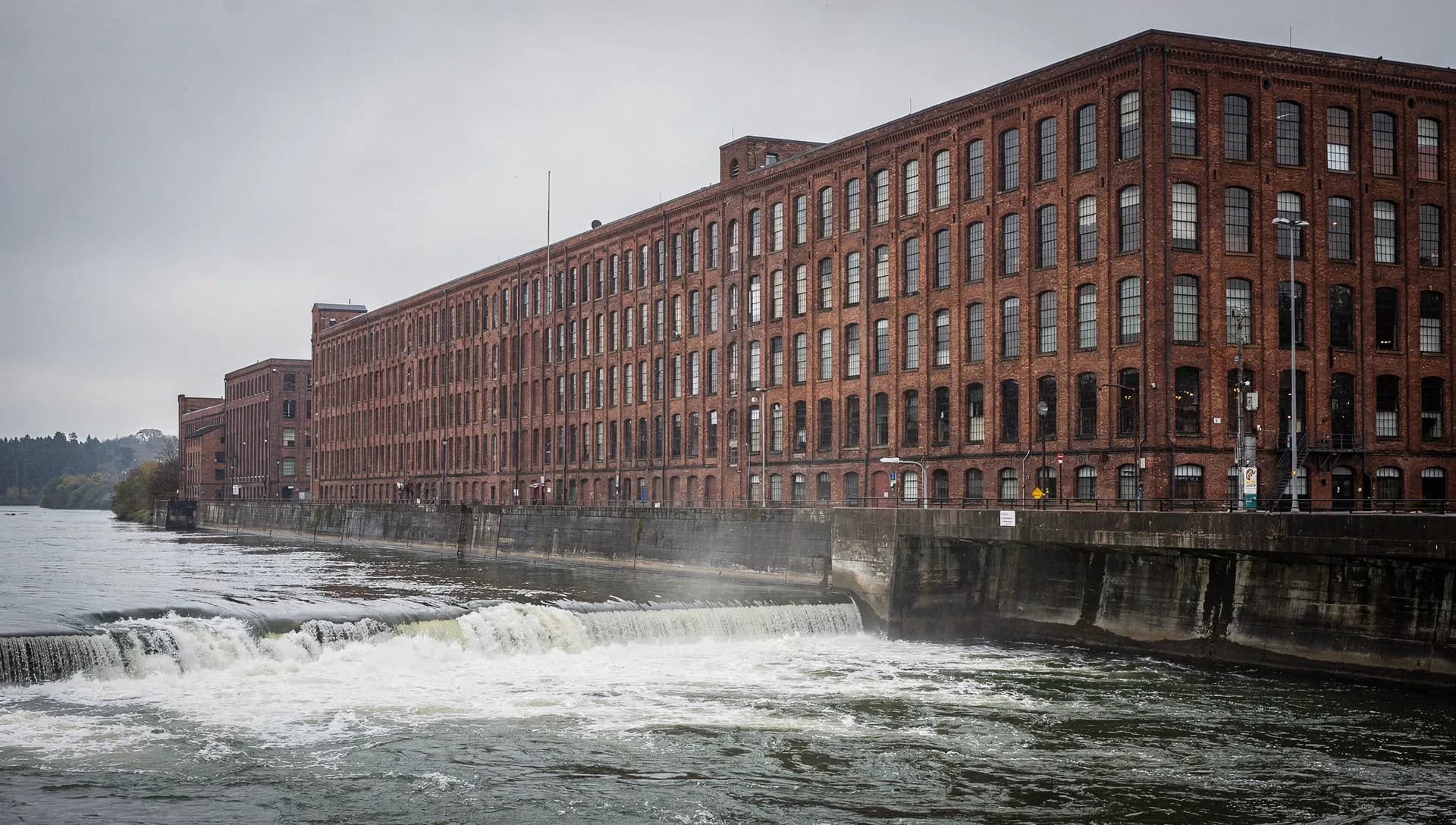 A long red-brick textile mill running along a river, rows of tall arched windows above a concrete embankment, a low dam spilling white water across the foreground under an overcast gray sky