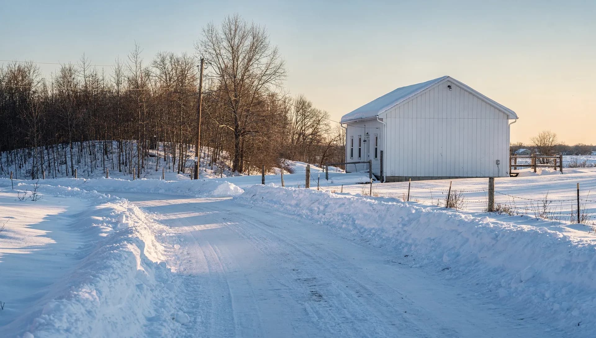 A plowed snow-covered country road at sunset leading past a white gable-roofed outbuilding with a snow-capped roof, a barbed-wire fence on wooden posts and bare trees under warm winter light