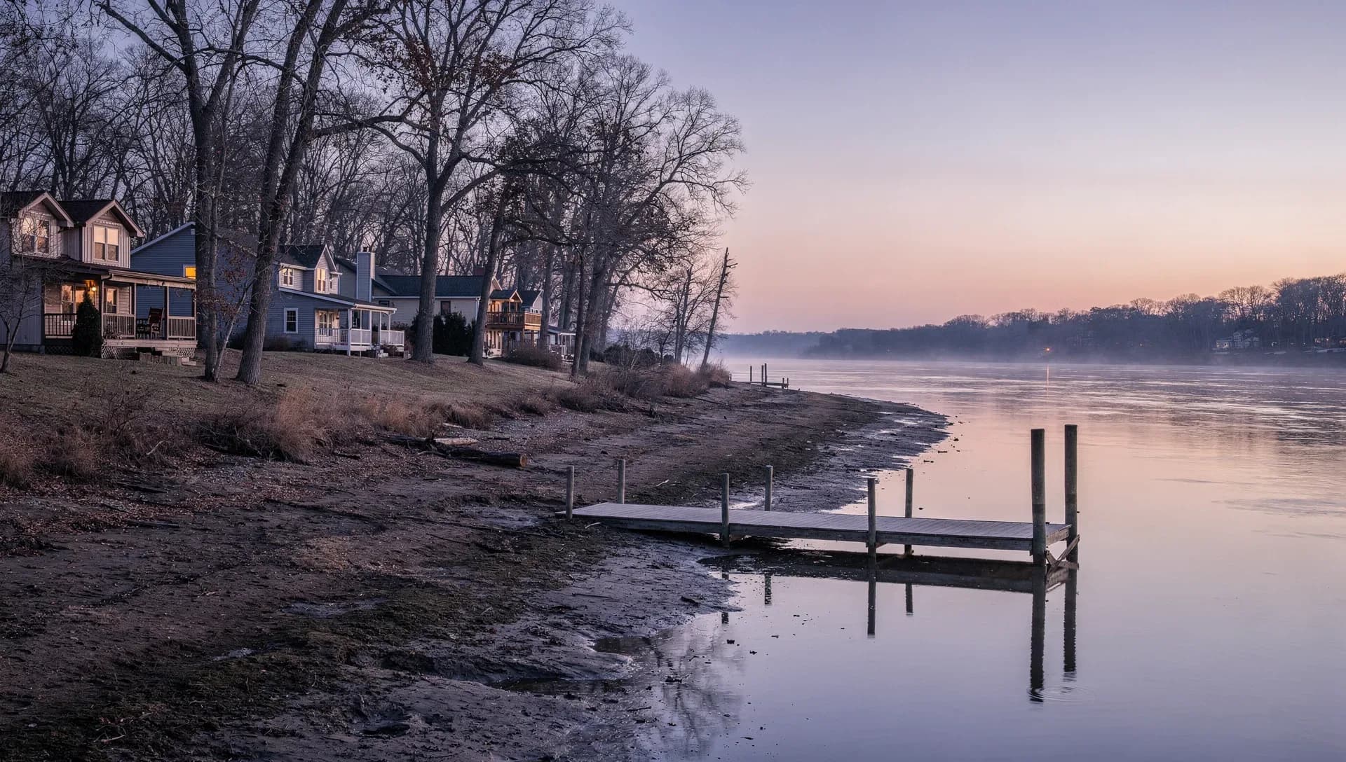 A calm tidal river at dusk with a small wooden dock on pilings reaching into still water, a row of houses among bare trees along a grassy bank, one window lit, a lilac and pink sky over the far shore