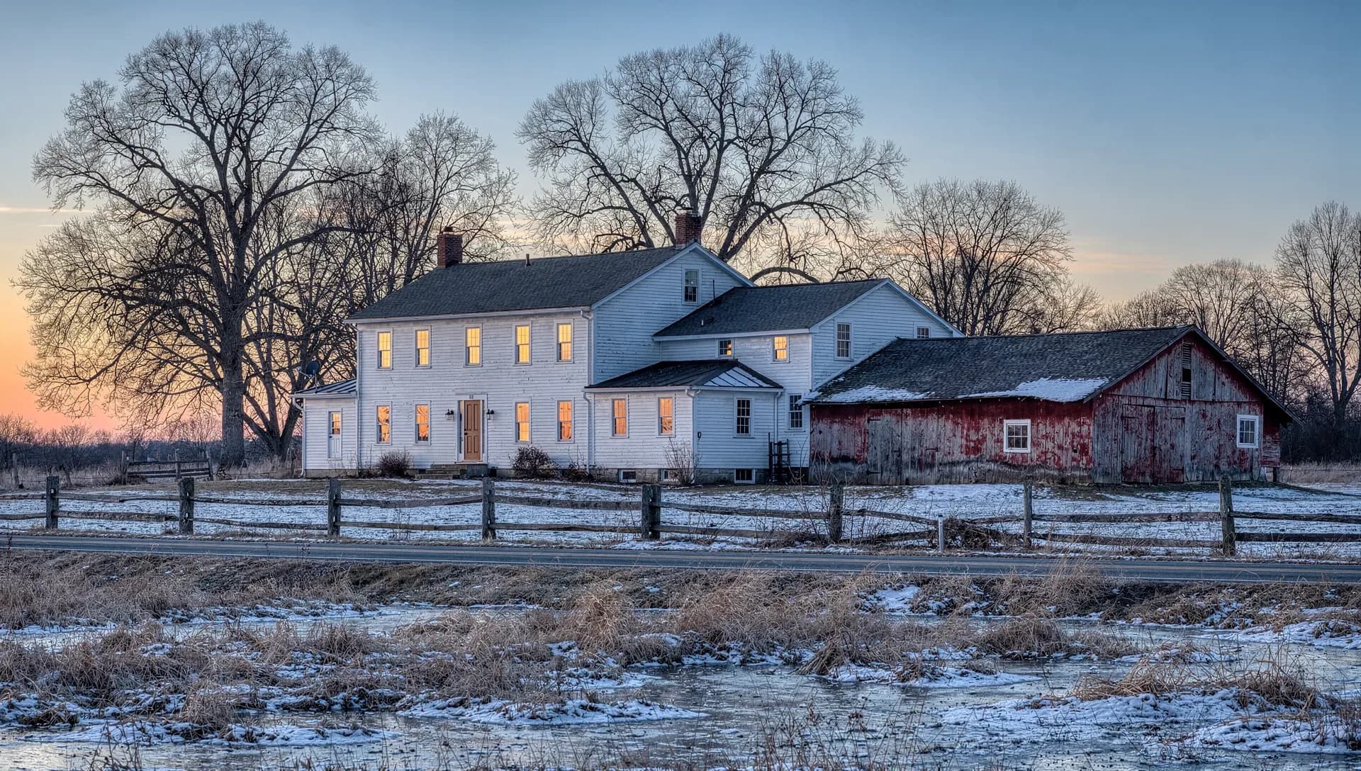 A white New England farmhouse with attached ells and a weathered red barn on a rural road, snow on the frozen fields under a dusk sky