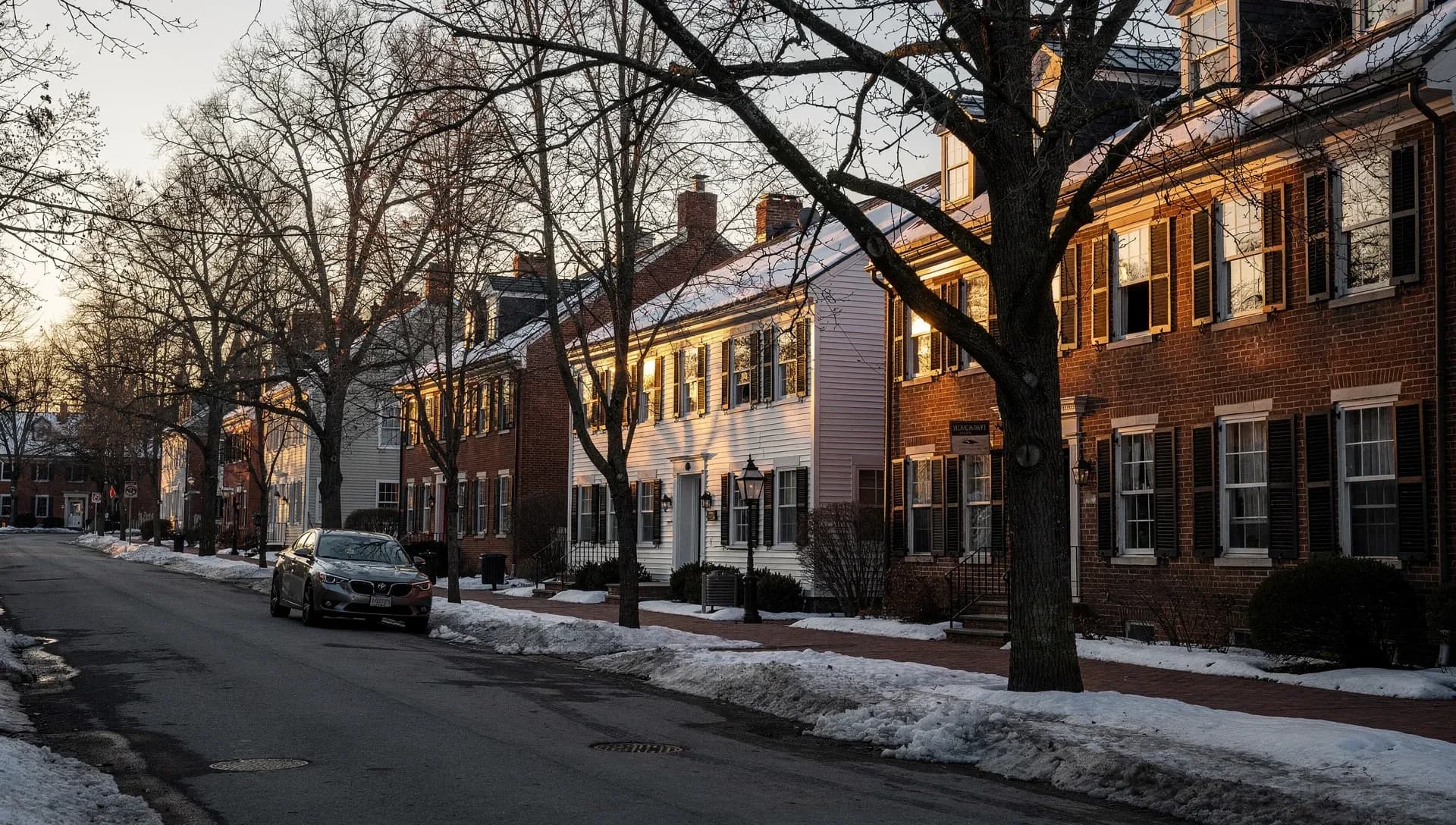 A row of brick and clapboard colonial houses along a tree-lined street in warm late-afternoon light, a car parked at the curb with snowbanks along the road