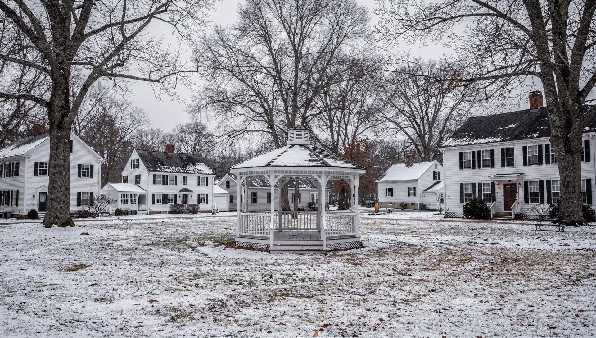 A white gazebo on a snow-dusted town common ringed by white colonial homes with black shutters and tall bare trees under an overcast winter sky