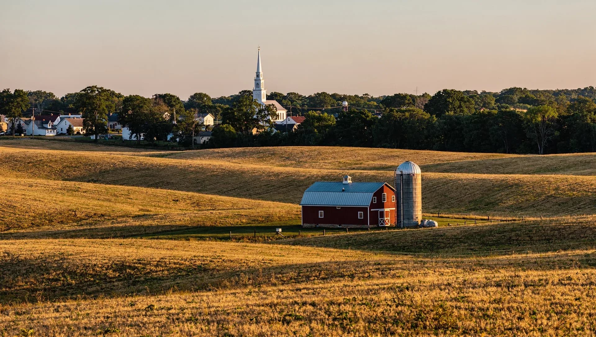 Rolling golden hayfields at sunset with a red gambrel barn and a metal silo in the foreground, a white church steeple and village rooftops set against a wooded treeline on the horizon