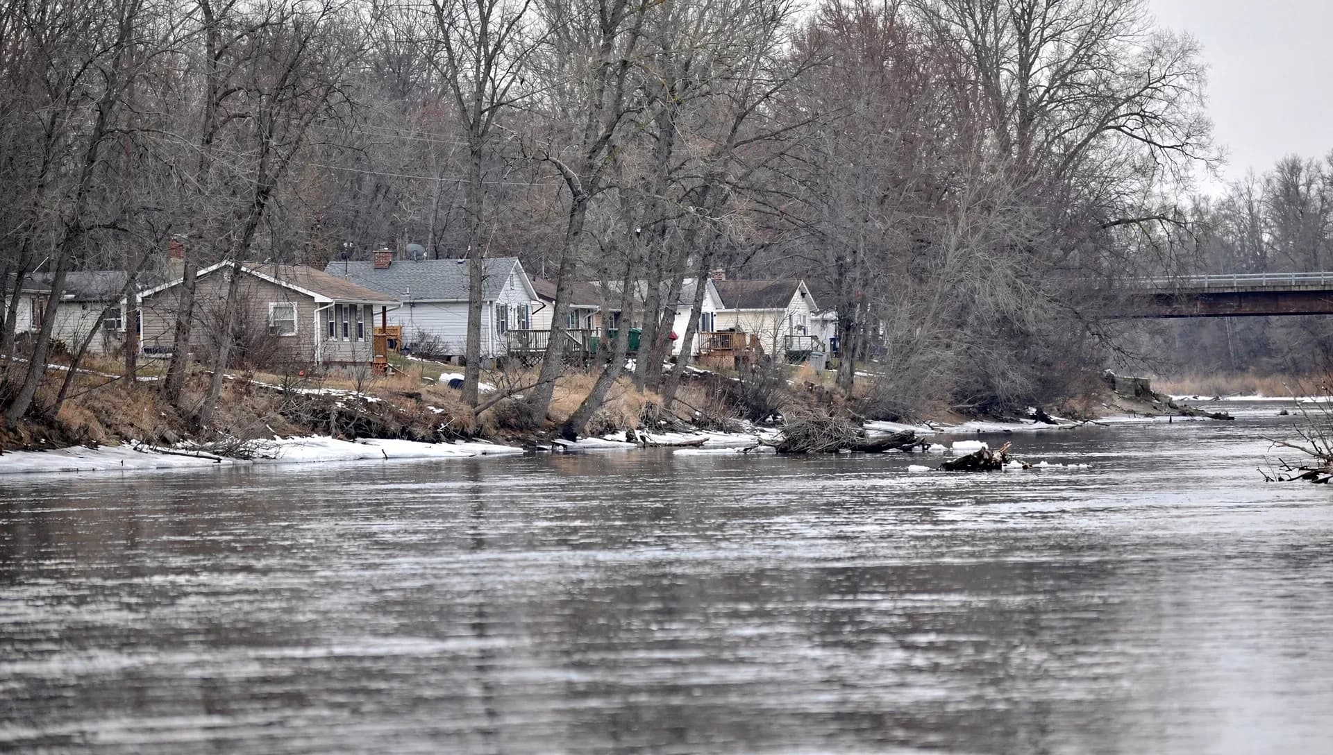 A wide grey river in winter with modest single-story houses and decks lining the far bank behind bare trees, patches of snow and ice along the shore and a low road bridge crossing on the right under an overcast sky