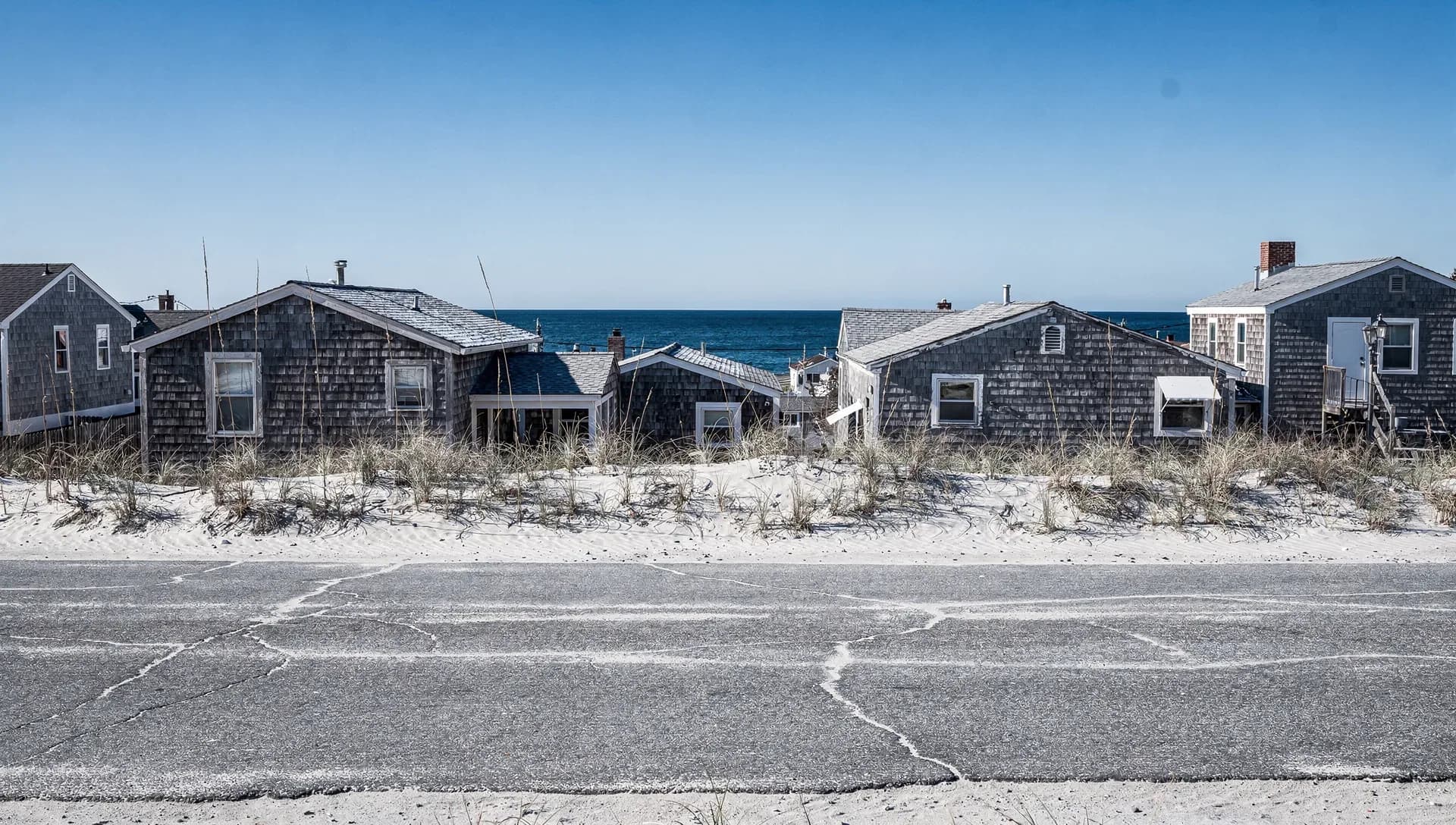 Weathered gray-shingled beach cottages behind dune grass with the open ocean beyond under a clear blue sky