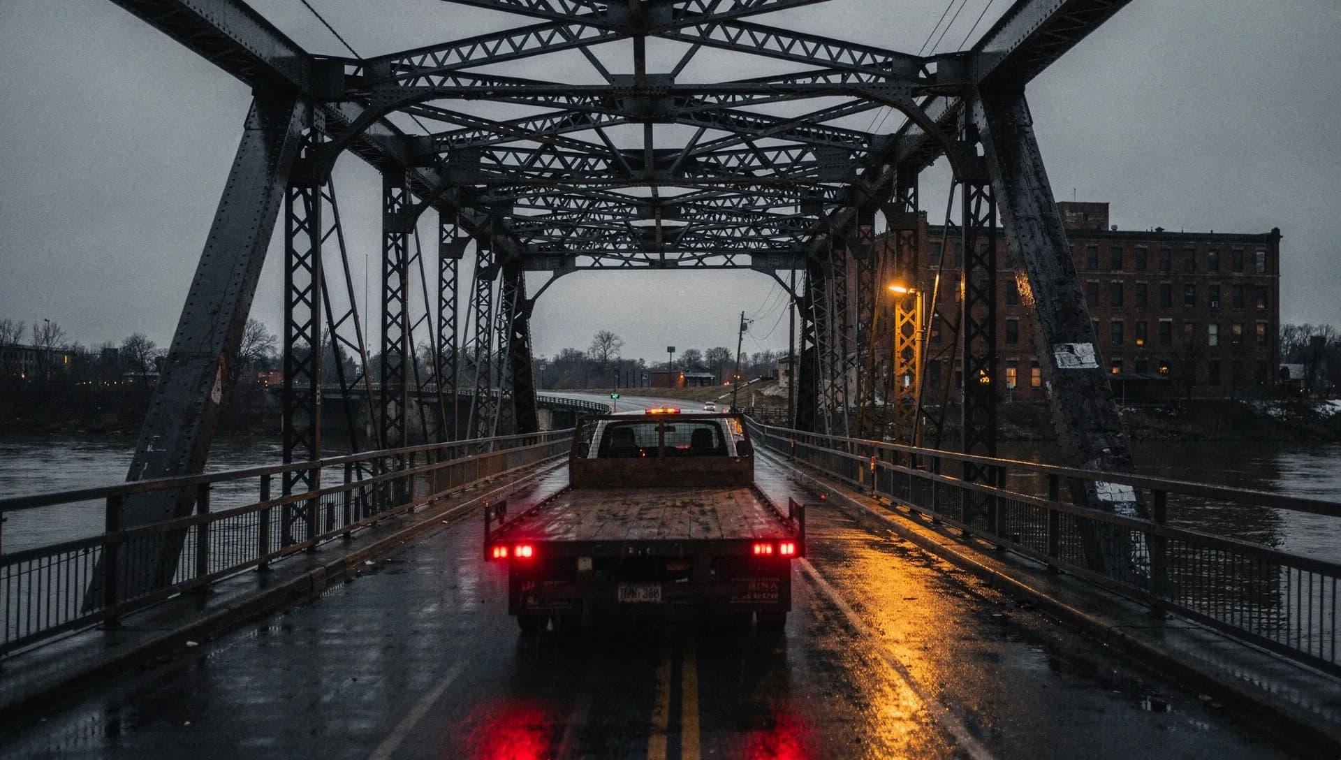 A flatbed work truck with its tail lights on crossing a riveted steel truss bridge in the rain at dusk, a brick mill building lit by one lamp on the far bank