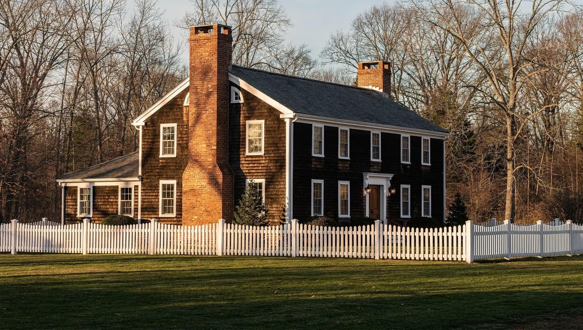 A large brown-shingled colonial house with two tall brick chimneys and white-trimmed windows standing behind a white picket fence and a mowed lawn, bare trees at golden hour