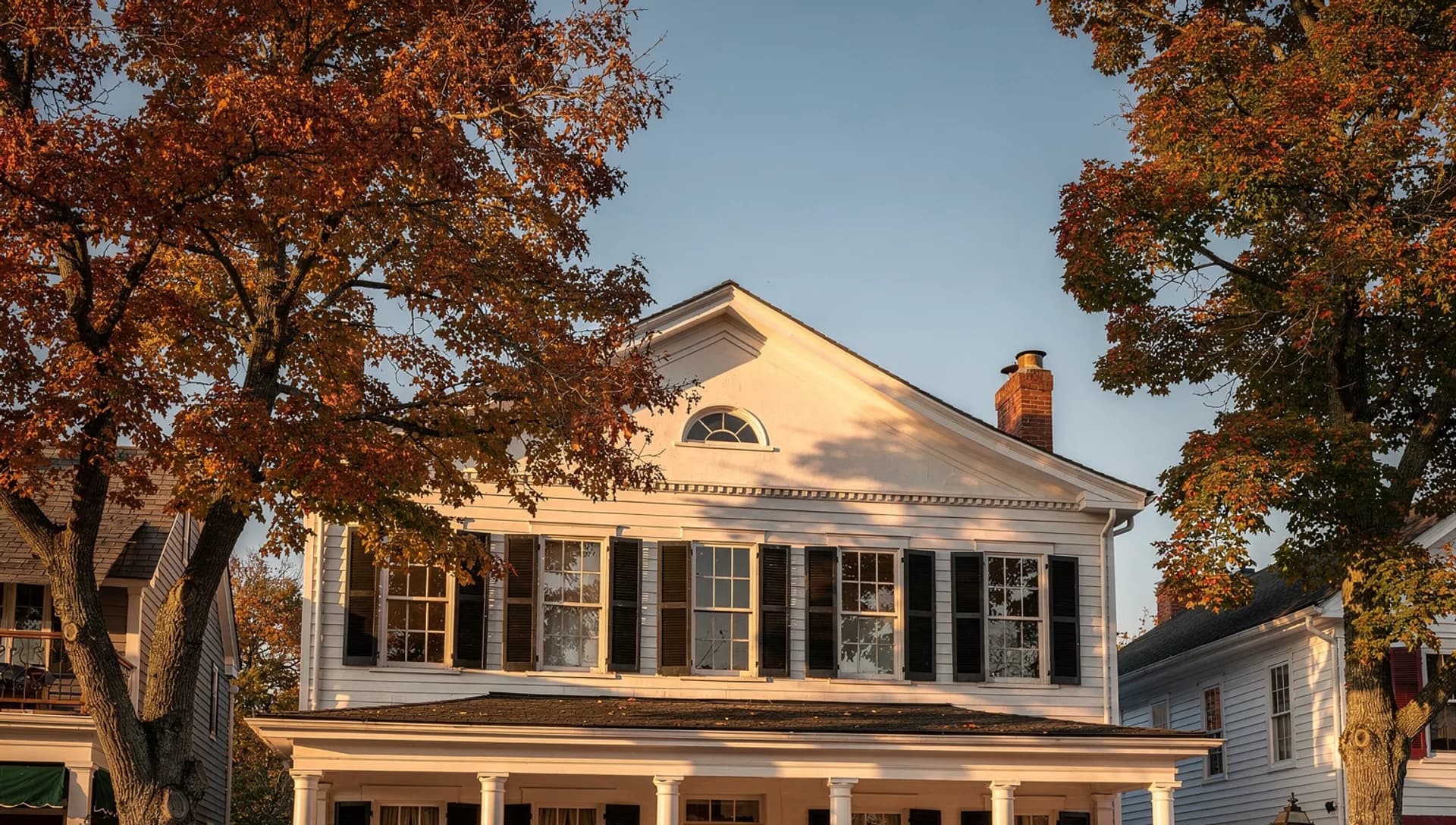 A white Greek Revival colonial with black shutters, a fanlight window set in its pediment, and a brick chimney, framed by tall oaks in autumn color against a clear blue sky at golden hour