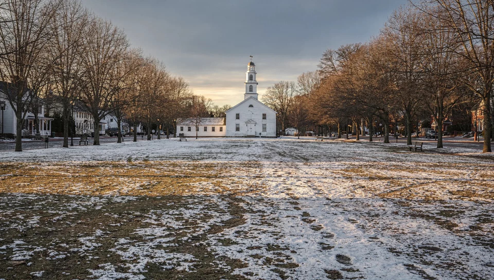 A white steepled meetinghouse at the head of a long snow-dusted town common lined with bare trees at sunset