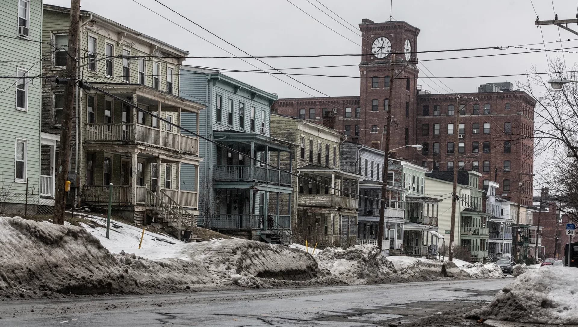 A row of tall wood-frame triple-decker houses with porches climbing a hill above a plowed, snowbanked street, a large brick mill with a prominent clock tower rising behind, overhead utility wires crossing an overcast sky