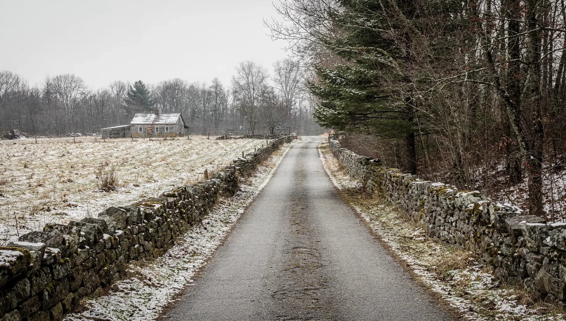 A narrow country road running between two long dry-stone walls dusted with snow, a weathered gray farmhouse with a metal roof in a bare field to one side, pines and leafless hardwoods under an overcast winter sky